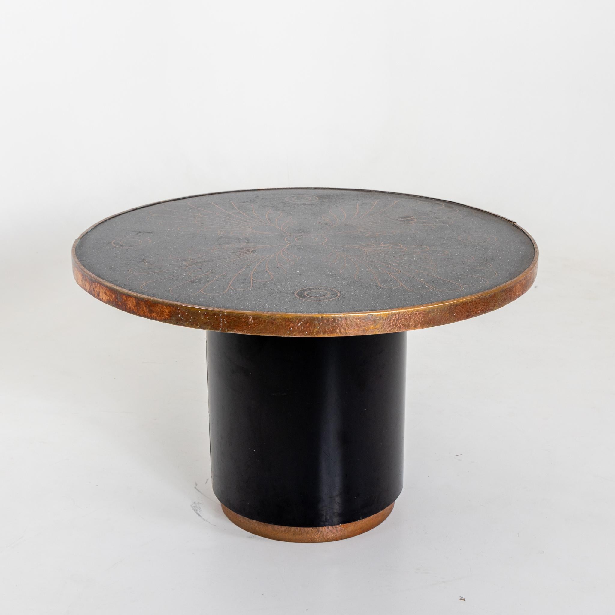 Large table with smooth round base and stone top with leaf-shaped metal inlays and hammered metal edge.