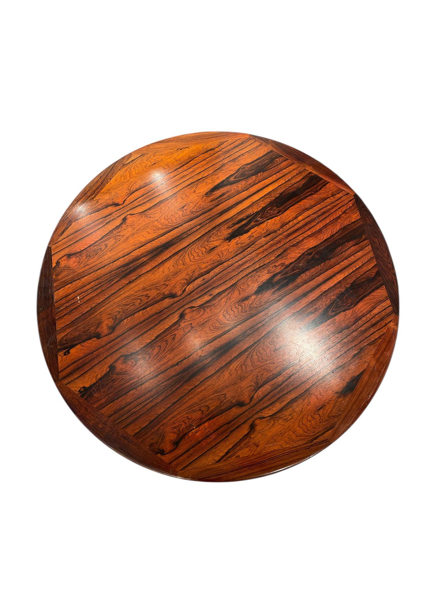 Una mesa redonda con tablero de madera oscura ricamente veteada. Se apoya en una base de metal cromado de cinco patas. El efecto general es de discreta elegancia, sofisticación y atemporalidad.