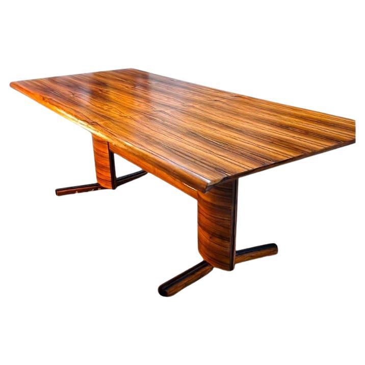 Questo tavolo da pranzo è caratterizzato da legno duro brasiliano con impiallacciatura Caviuna. Vanta un piano rettangolare con bordi arrotondati, sostenuto da due gambe collegate da una trave anch'essa con bordi arrotondati. Il legno è stato