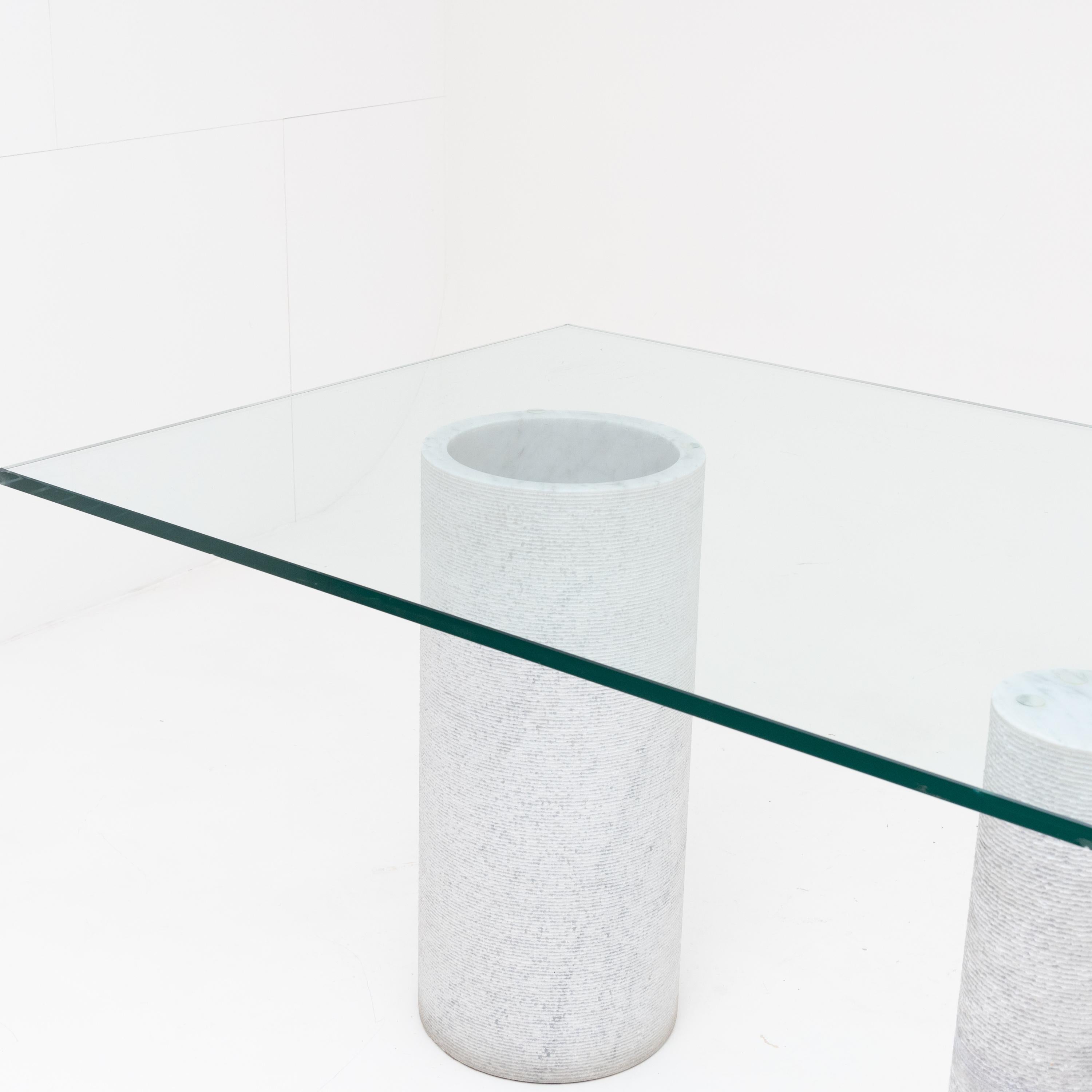 Large dining table with glass top on three cylindrical marble bases of different thicknesses with grooved surfaces.