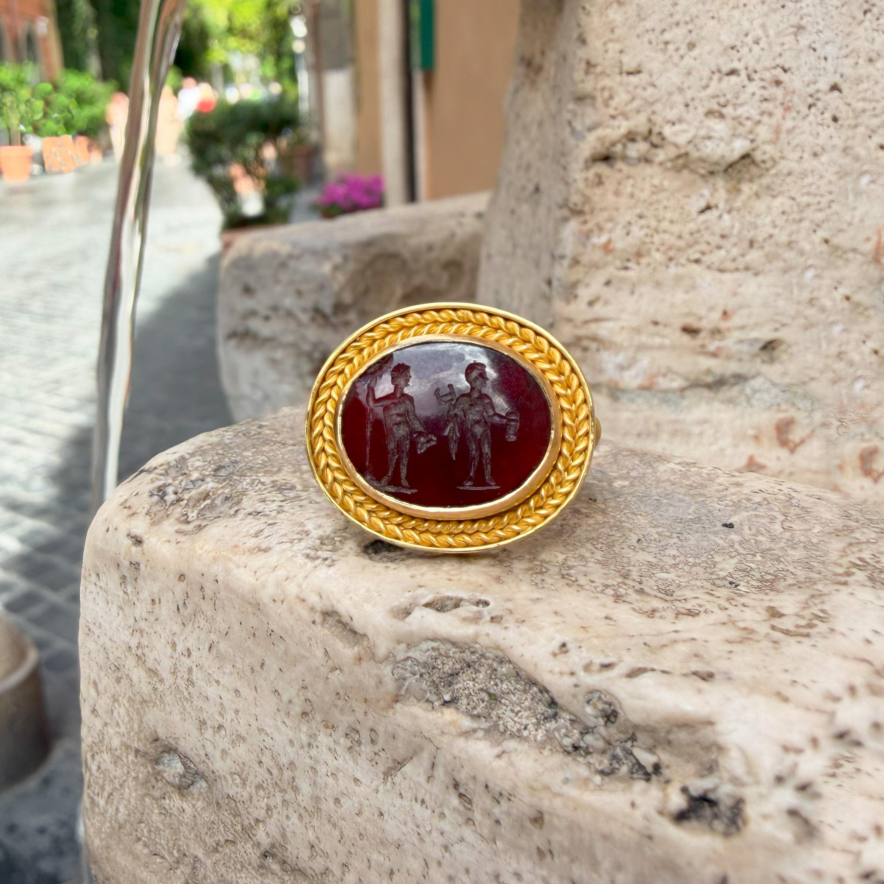 Cette bague, fabriquée à la main par nos orfèvres en or 22 kt, présente une authentique pierre de cornaline du 1er-2e siècle ADS, représentant deux figures masculines : Dionysos à gauche et Apollo à droite.
Dans sa main droite, Dionysos tient un