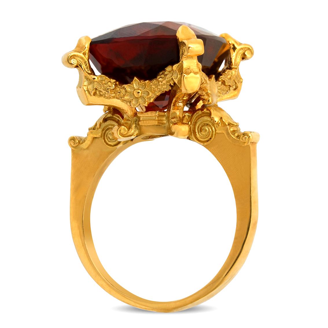 Handcrafted in 18kt yellow gold this lavish ring features a deep wine coloured central 20.98ct cushion cut garnet aloft a signature William Llewellyn Griffiths garland setting resplendent amongst intricate flowers, baroque scrollwork and cross