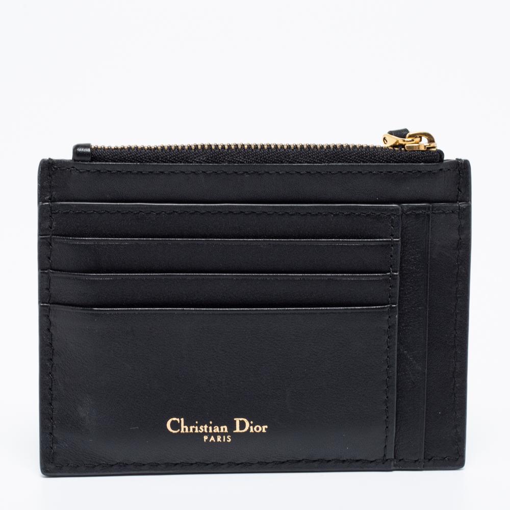 The brand signature in gold tone adorns the black exterior of this Dior card holder. Created from leather, the design is highlighted with a compartmentalized front and features a zipper closure at the top. The fabric-lined interior can neatly keep
