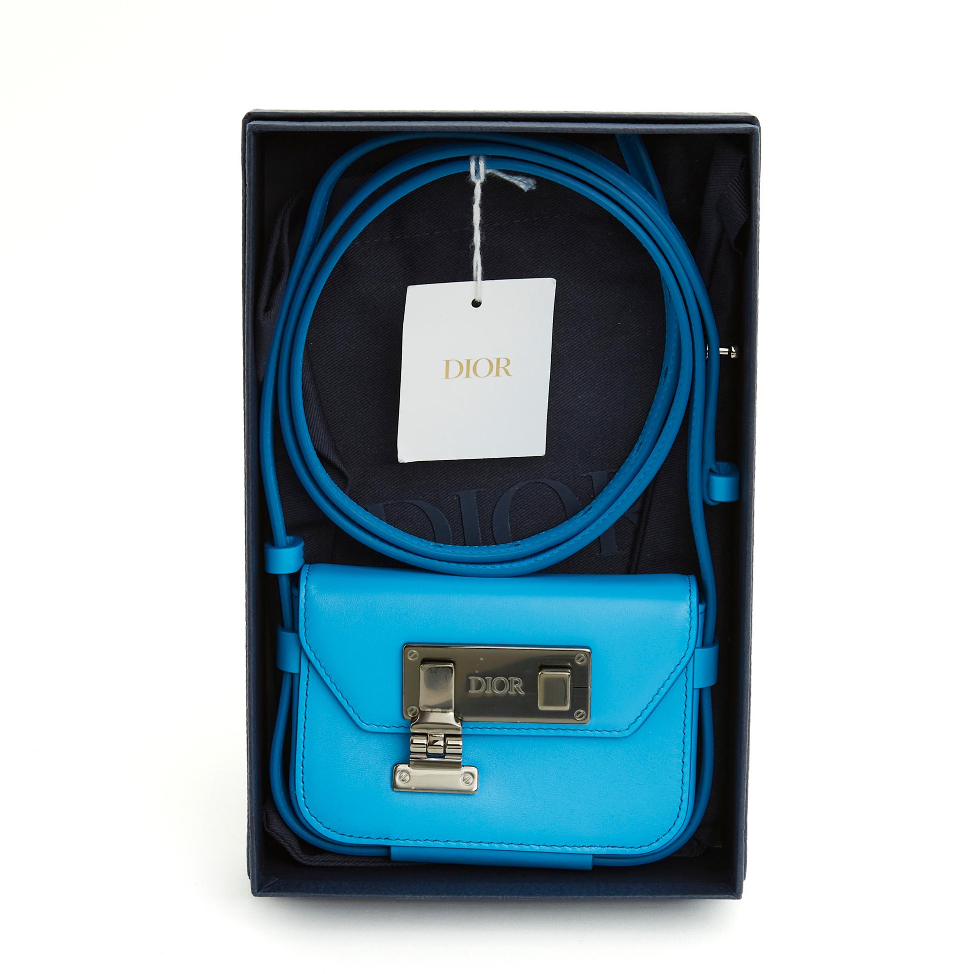 Pochette Nano Dior en cuir bleu roi, fermoir en métal argenté légèrement foncé (ruthénium) avec le logo Dior, intérieur en toile noire avec une pochette plaquée pour une carte, longue bandoulière réglable pour un portage croisé. Largeur 11,5 cm x