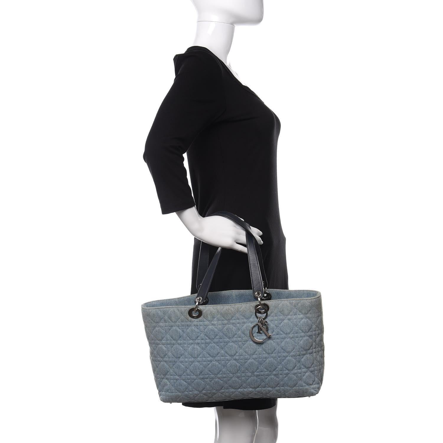 This Dior bag from 2005, designed by John Galliano, is constructed of blue denim with a signature Cannage pattern. It features silver-tone hardware, silver Dior charms, dual shoulder straps, and leather trim details. The open-top design reveals a