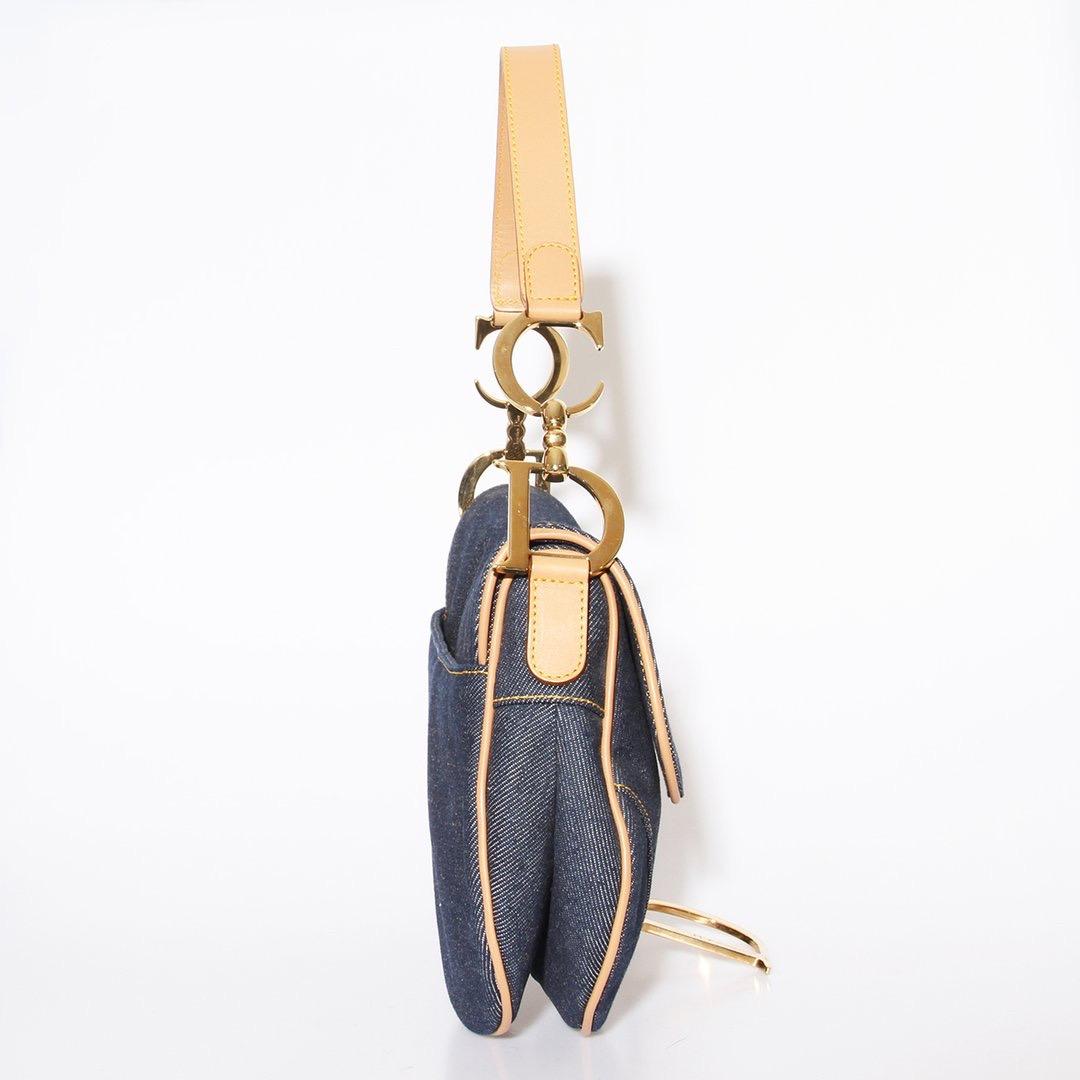 Denim saddle bag by Dior 
Dark blue denim 
Tan leather trim 
Velcro closure 
Gold tone metal hardware
Features a top handle adorned with metal 'CD' finishing
Saddle-shaped silhouette
Interior zip pocket 
Patterned interior lining 
Authentication