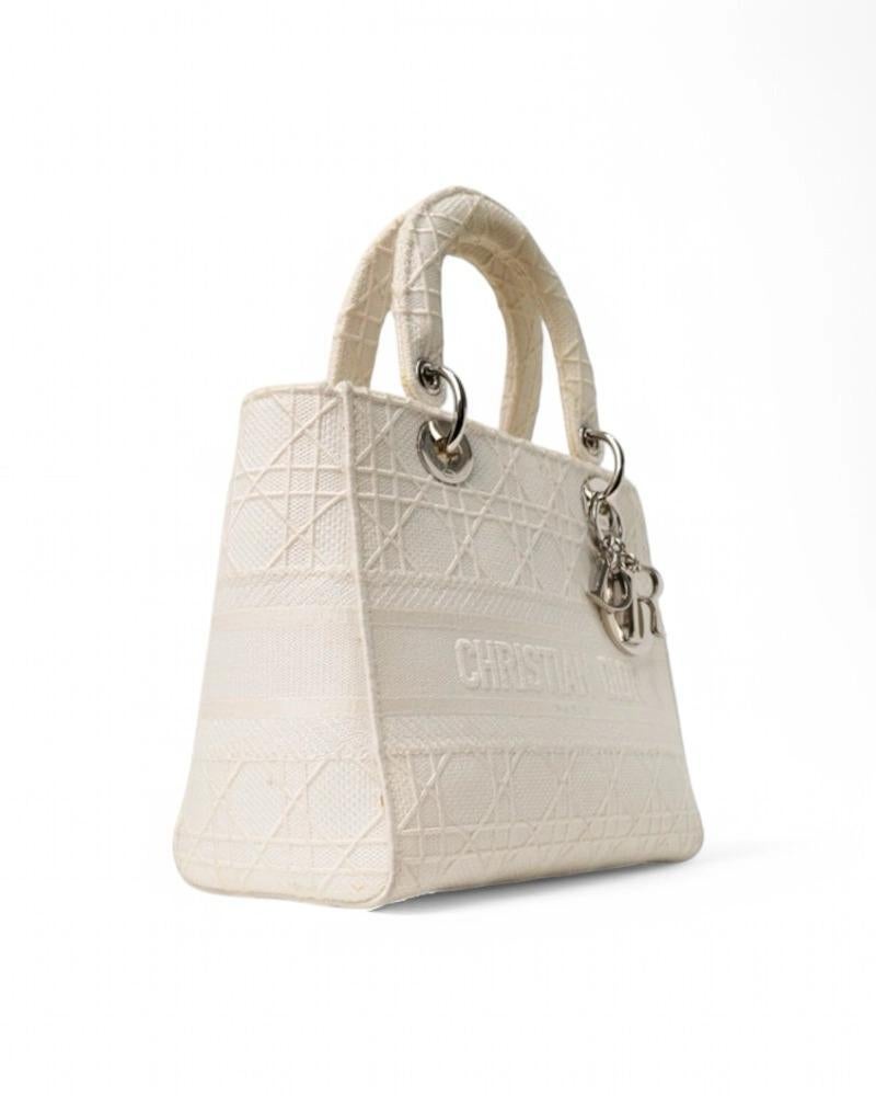 Sac de créateur Dior, modèle Lady D-Lite en taille Medium, en tissu crème avec motif cannage et ferrures argentées. Doté d'une fermeture à rabat et doublé à l'intérieur en tissu ton sur ton avec une poche pratique, le sac est assez spacieux. Équipé