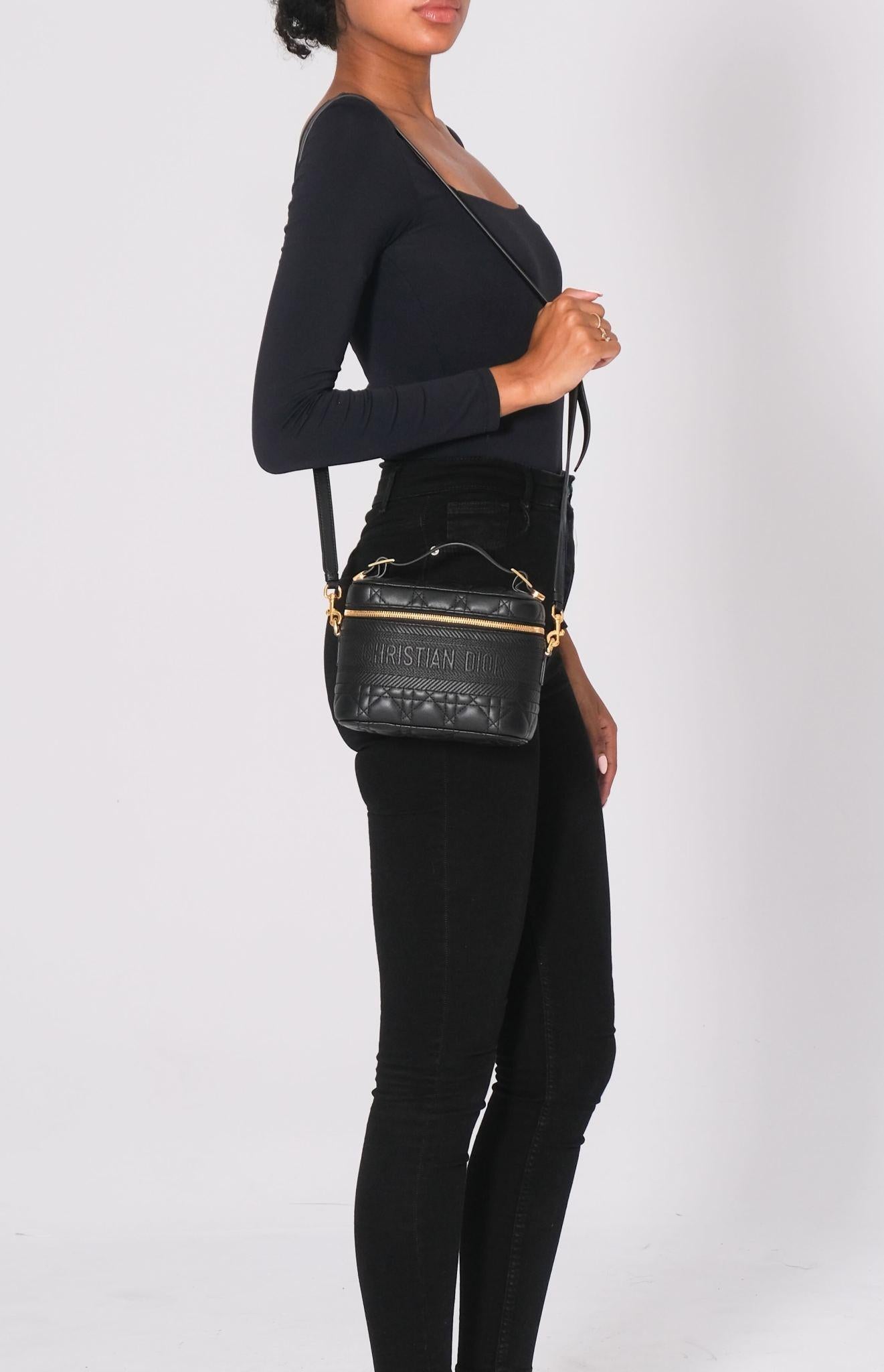 Ce vanity case Dior est réalisé en cuir d'agneau souple et doux, de couleur noire, avec les piqûres de cannage caractéristiques de la marque. Le sac est orné d'un logo Christian Dior sur le devant, d'une anse supérieure et de ferrures dorées. La