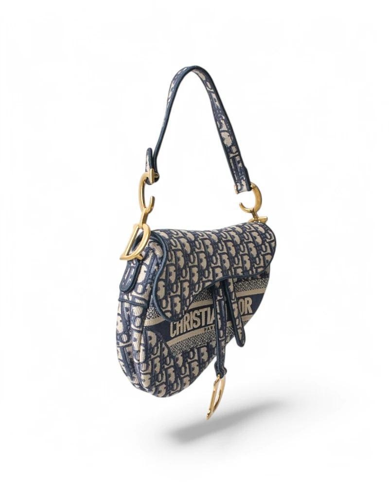 Bolso de Dior, modelo Saddle. Realizado en lona azul con el clásico motivo jacquard Dior Oblique, está acabado en piel lisa y herrajes dorados. La solapa delantera tiene un cierre magnético en forma de 