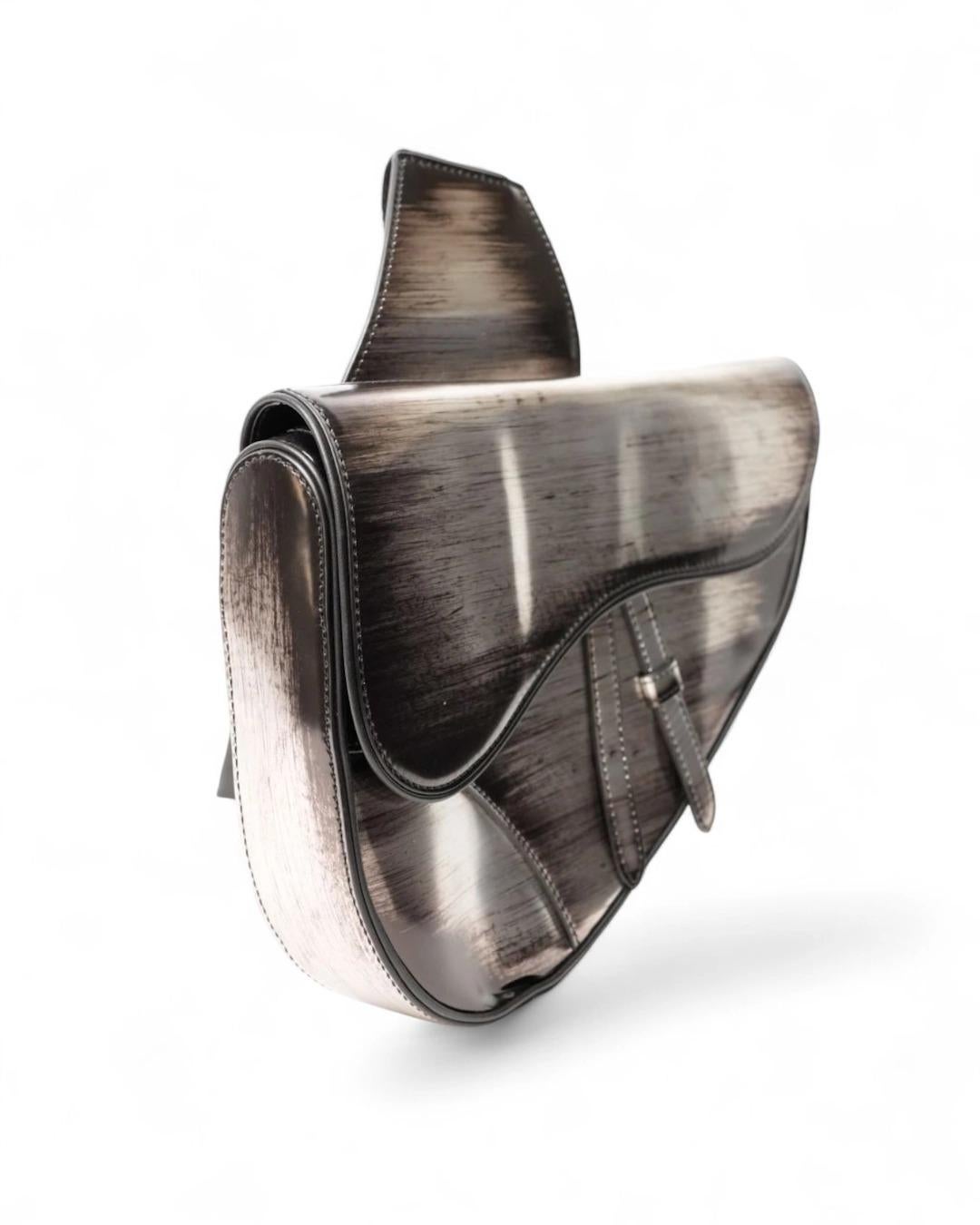 Bolso Dior Saddle de piel plateada brillante con motivo degradé y herrajes plateados, solapa delantera con botón magnético y cierre interior de cremallera. Forrado internamente en lona negra, el bolso es espacioso para lo esencial. Equipada con una