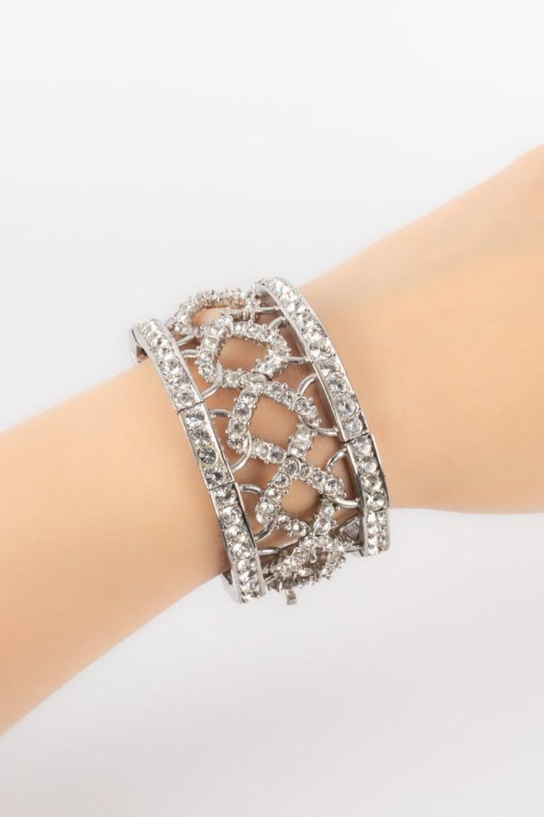 Dior - (Fabriqué en France) Bracelet articulé en métal argenté orné de strass, d'un nœud et des initiales de la marque.

Informations complémentaires : 
Condit : Très bon état.
Dimensions : Circonférence : 17 cm - Largeur : 3 cm

Référence du