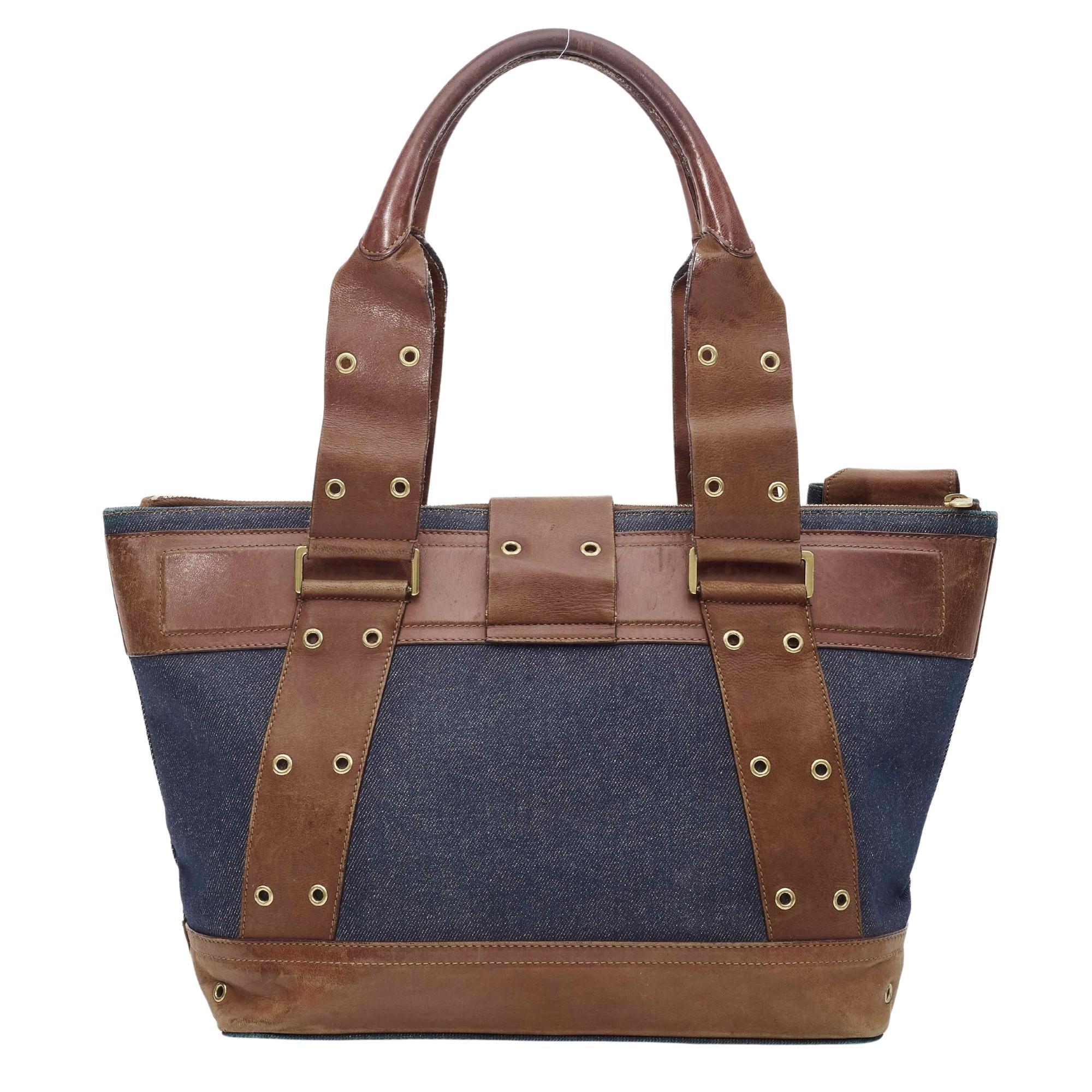 This shoulder bag is made of fine blue aged denim and features brown leather trim, dual rolled top handles, brass hardware and a a crossover strap with a button. The top zipper opened to a brown Dior logo fabric interior with a zipper pocket.