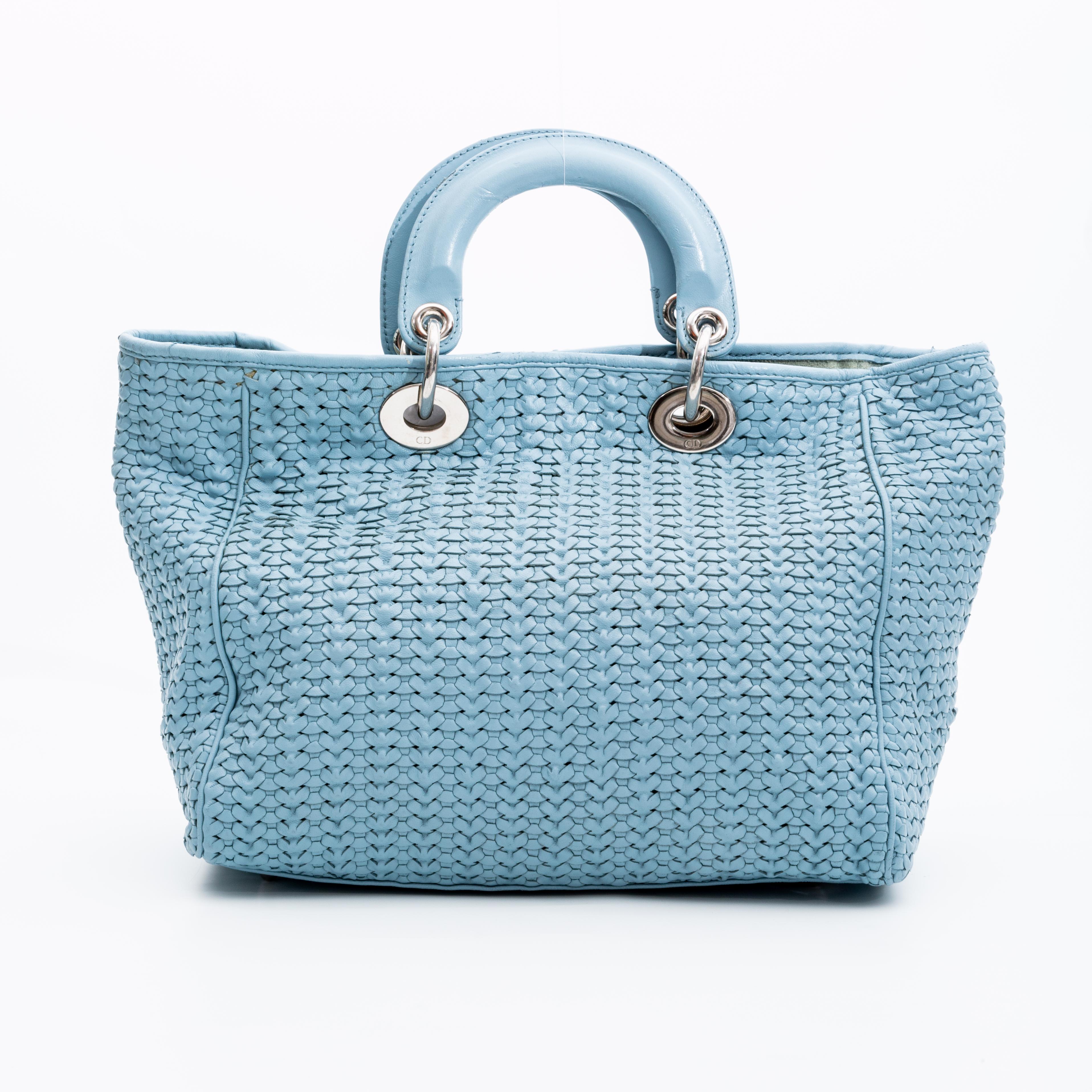 Ce sac iconique Lady Dior est le modèle de petite taille et présente du cuir tressé bleu pastel, des ferrures de couleur Icone, des charmes Dior, deux anses attachées par des liens ronds, un dessus ouvert avec fermeture à bouton-pression et une