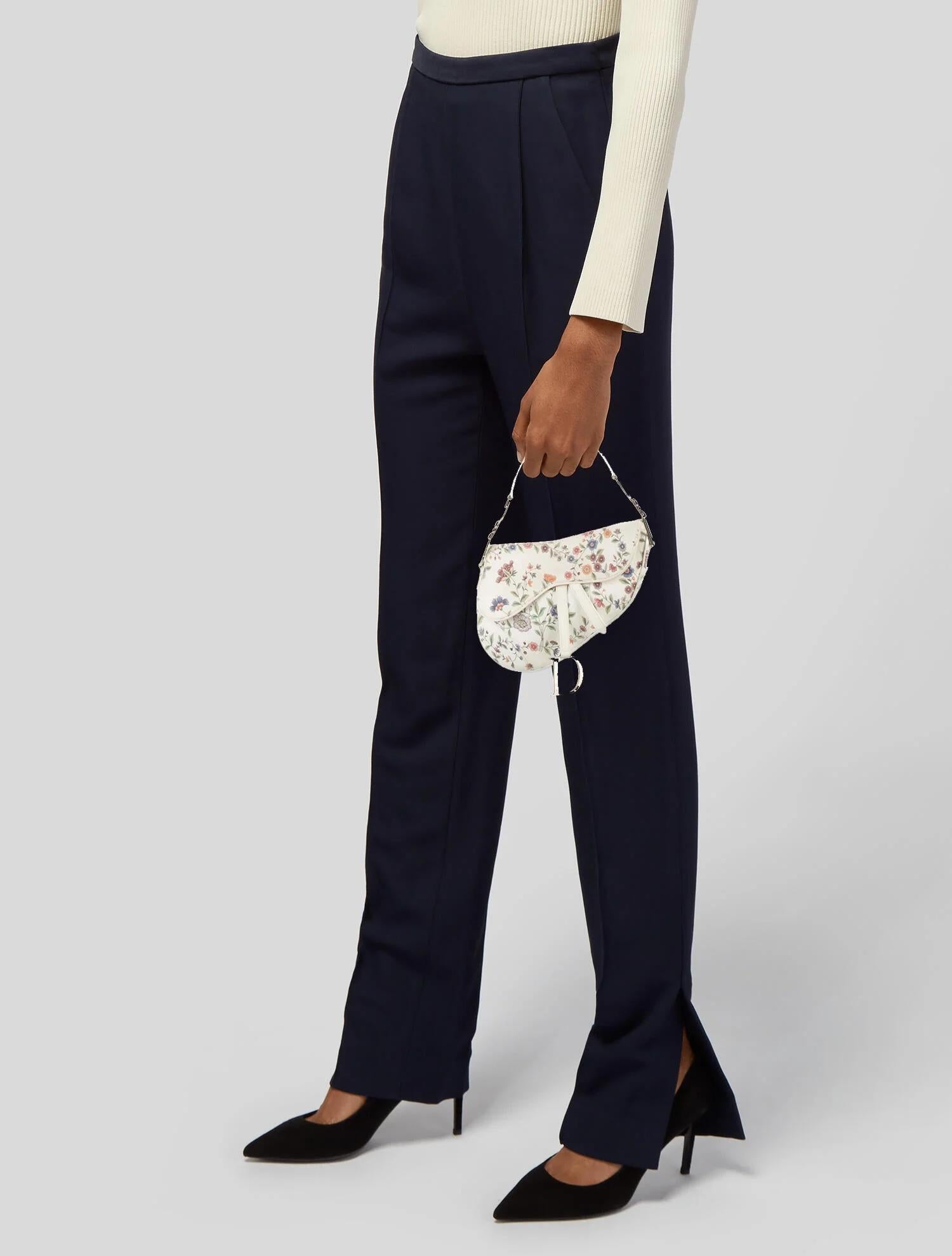 This Limited Edition Dior mini saddle bag is made of white canvas with a multicoloured floral print. The bag features a leather shoulder strap with logo charms, silver hardware, and a front flap. The front flap opens to a Dior oblique jacquard cloth