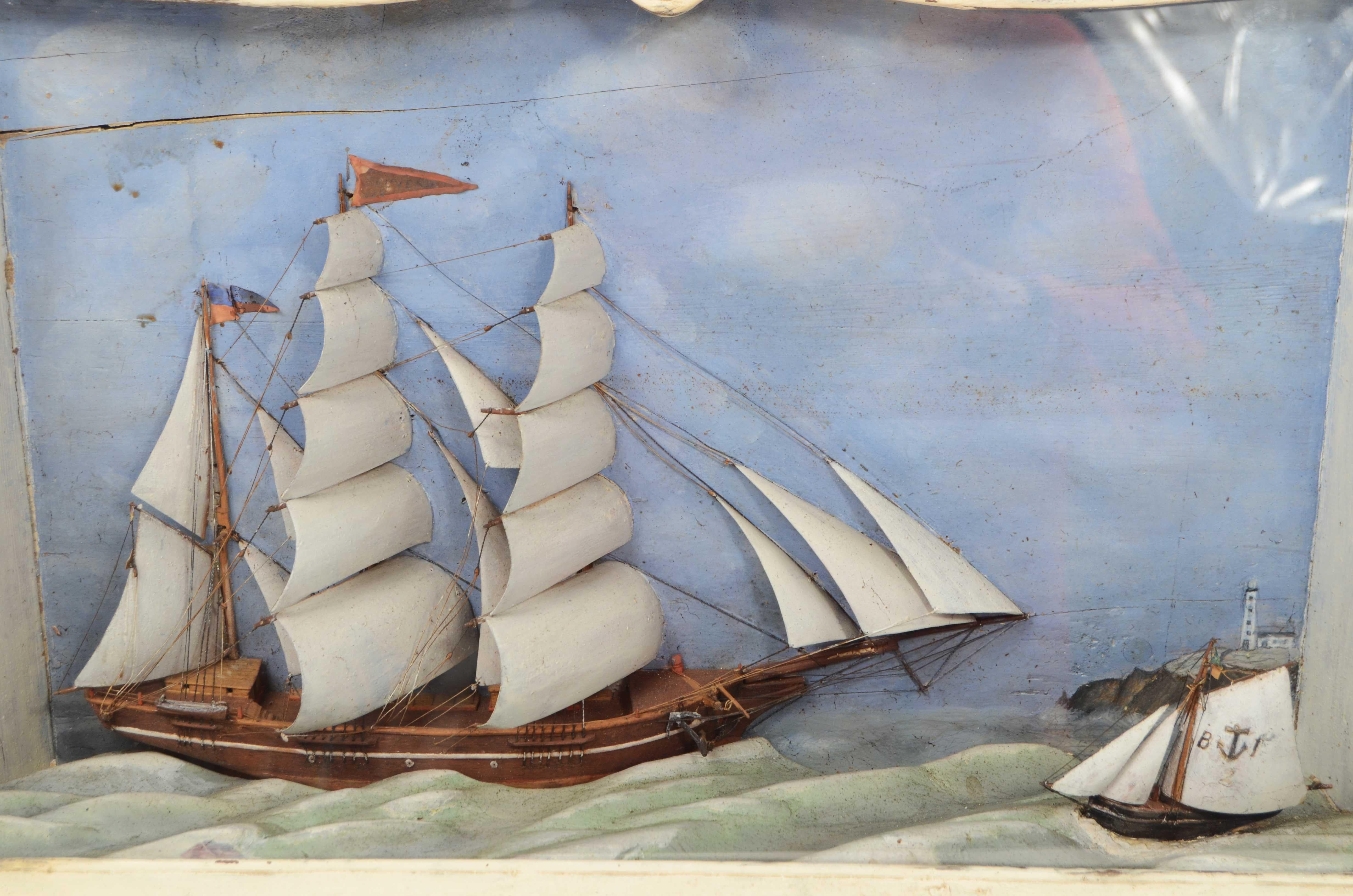 Diorama nautique dans un coffret trapézoïdal en verre et bois peint en blanc représentant la demi-coque dans les vagues d'un brigantin aux voiles déployées en balsa, à l'arrière-plan un promontoire avec un phare peint et sur le côté un autre bateau