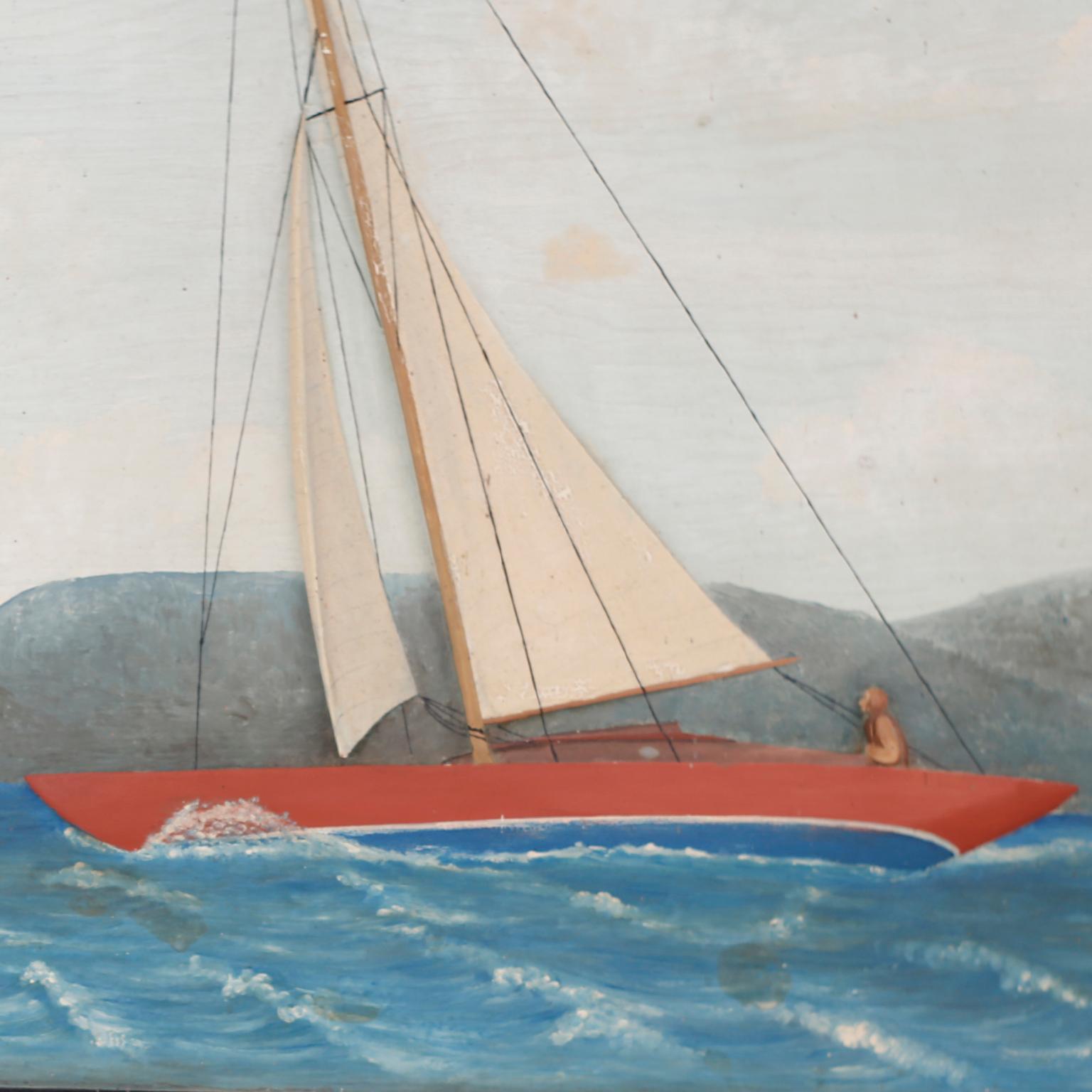 Vintage diorama of a red sailboat on a choppy lake carved and painted on a wood panel in a naive folk style, presented in a wood frame decorated with anchors and titled Finnan Haddie and dated 1933.