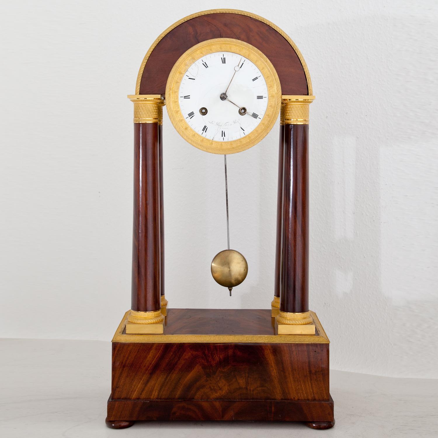 Mahogany-veneered Directoire portal clock, standing on a rectangular base. Four columns with gilt bases and capitals support the clockwork, which is housed in a cylindric brass case. The molding and the ring around the enamel clockface is gilt as