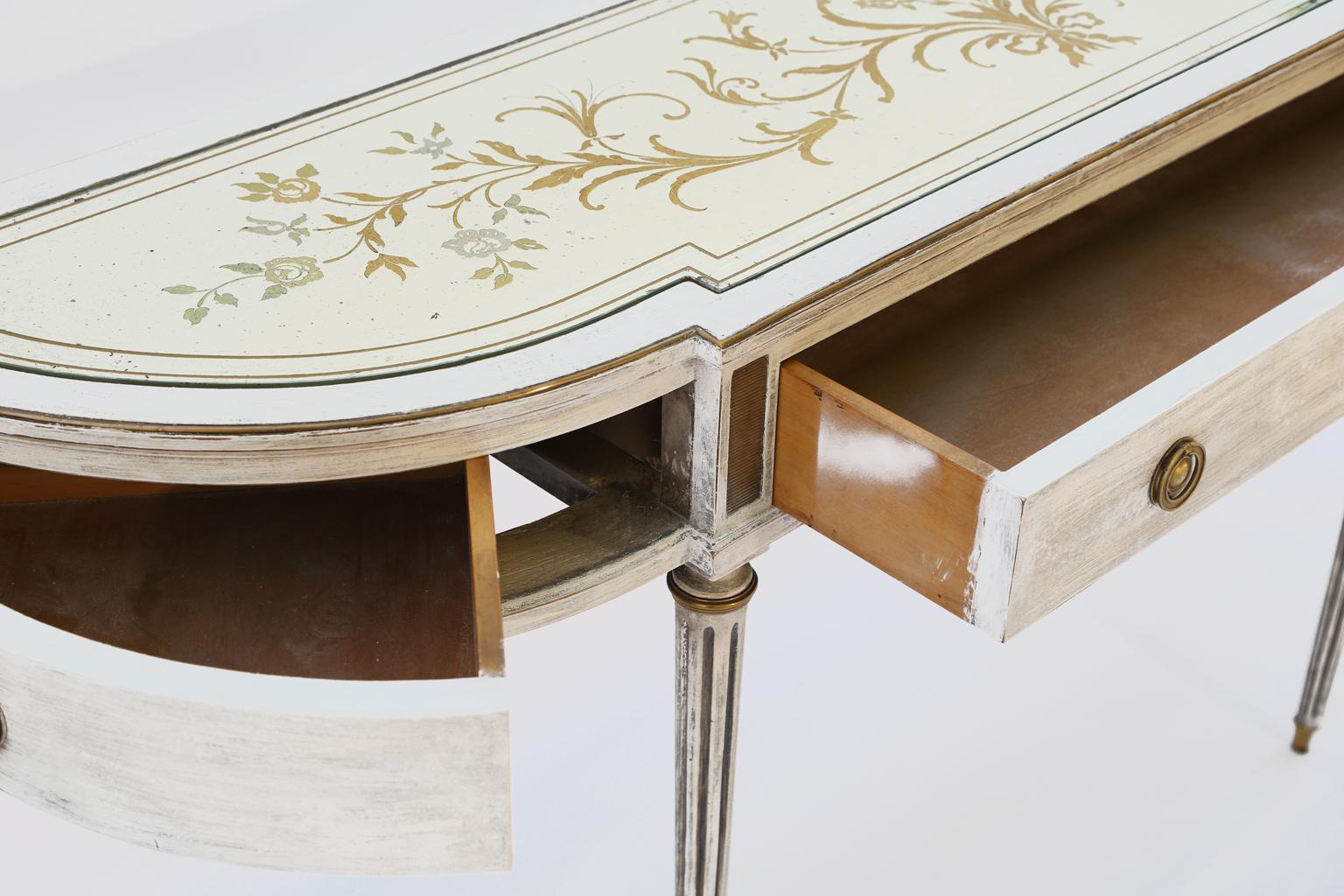 Demilune console table, in Directoire taste, of mahogany, with a painted finish showing natural wear, its églomisé top etched and reverse painted in gold and silver scrolling floral motif, a brass trim borders its top and legs, triple frieze drawers