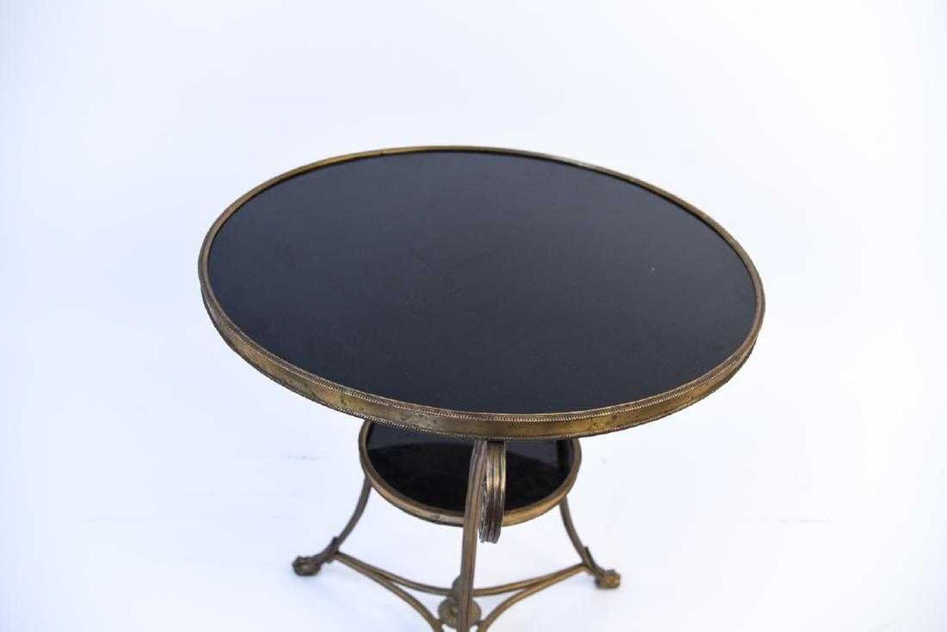 A French mid-20th century Directoire style guéridon brass table with stone top and lower tier. The table is raised on three saber-shaped legs, beautifully scrolled in their upper section and resting on lion paw feet. The legs are connected to one