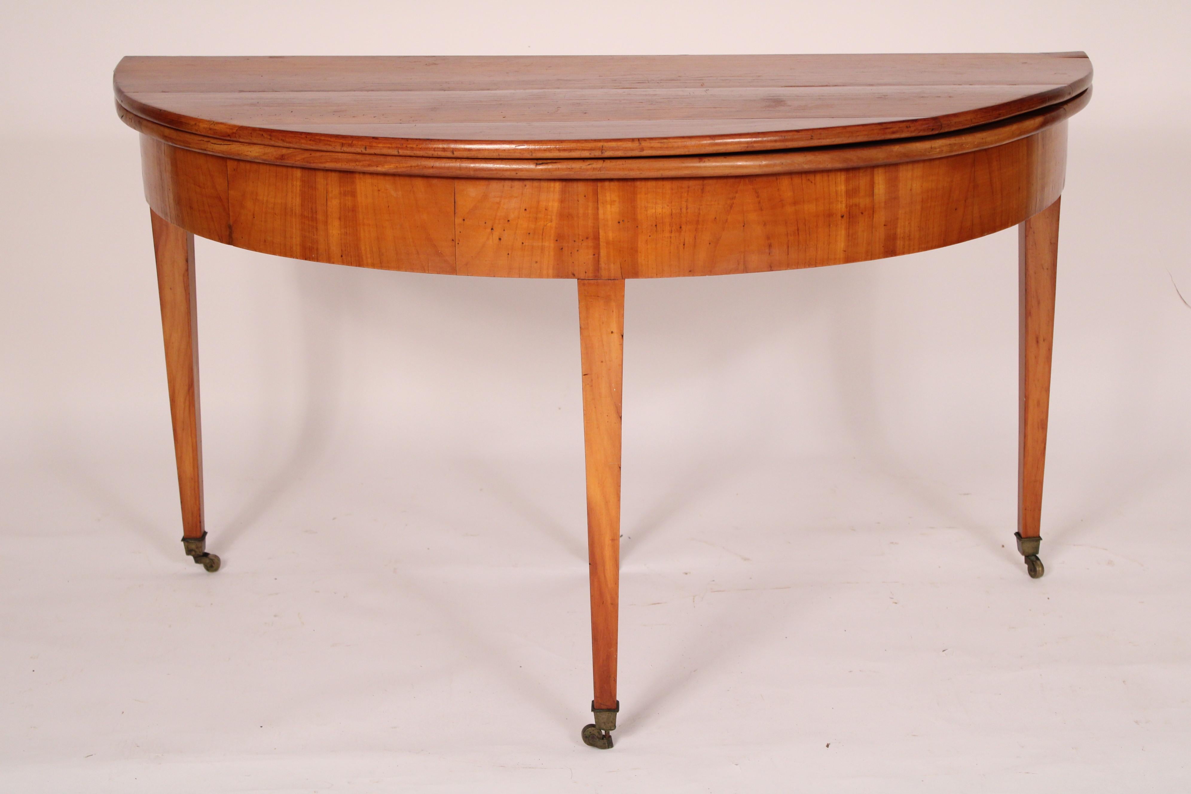 Continental Directoire fruit wood demi lune games table, early 19th century. Having a demi lune top which when opened becomes a round top, a fruit wood frieze, resting on square tapered legs ending in brass casters.  Knee clearance 23.25