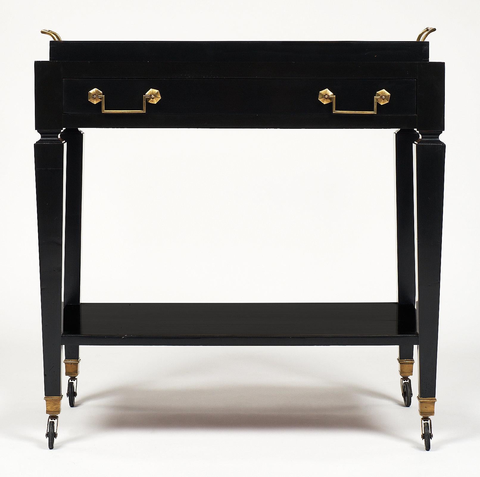 An elegant French Directoire style tray table on casters with a detachable tray featuring brass handles and a glass bottom. This piece is made of ebonized cherry wood. The “service” bar cart also has a dovetailed drawer with two handles and a bottom