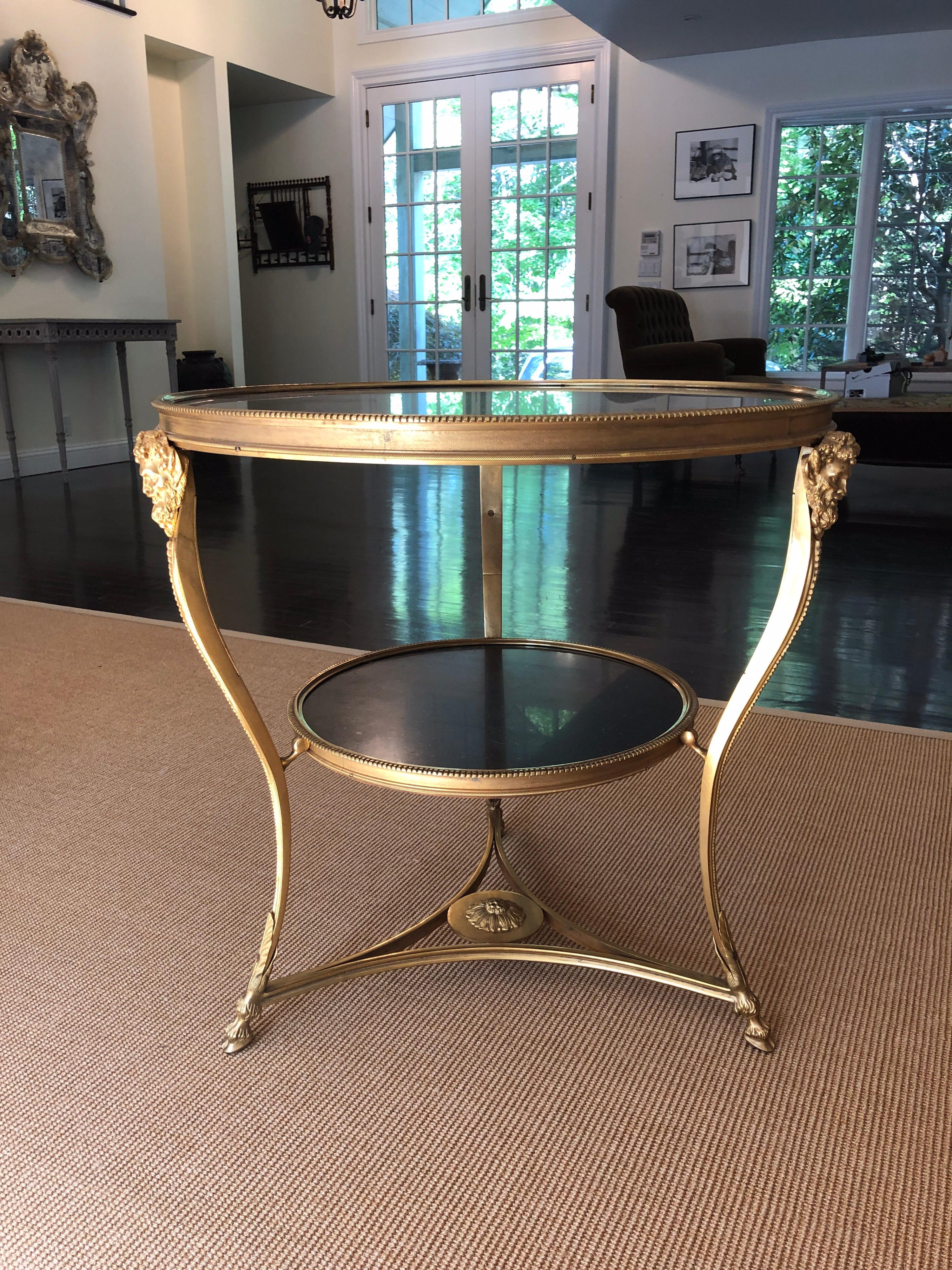 Directoire style gilt bronze gueridon table with dark marble top. Two tiers with dark marble tops, gilt bronze Bacchus heads, ram hoof feet and central medallion on lower structure. This is in excellent condition.
