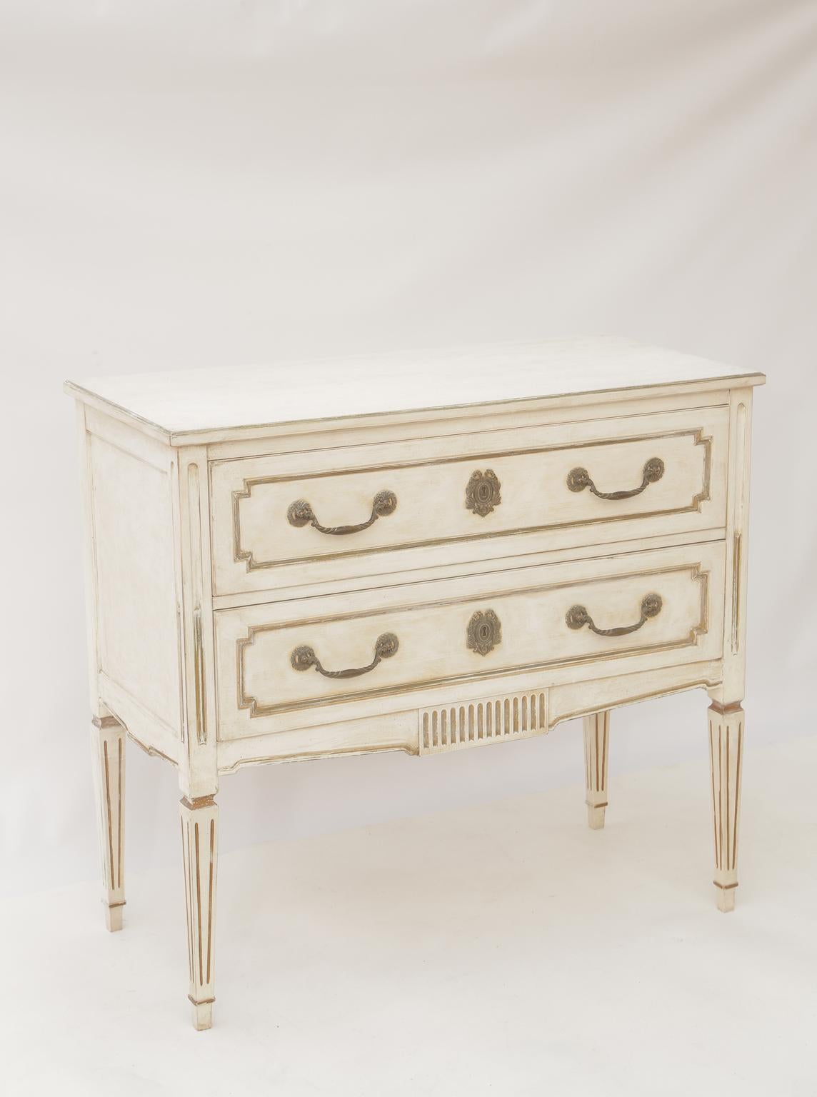 Vintage commode, by Henradon, in Directoire taste, having a painted and parcel-gilt finish showing natural wear, its rectangular top on a base of two stacked drawers with double pulls, centered by an escutcheon, framed by moldings, over a fluted