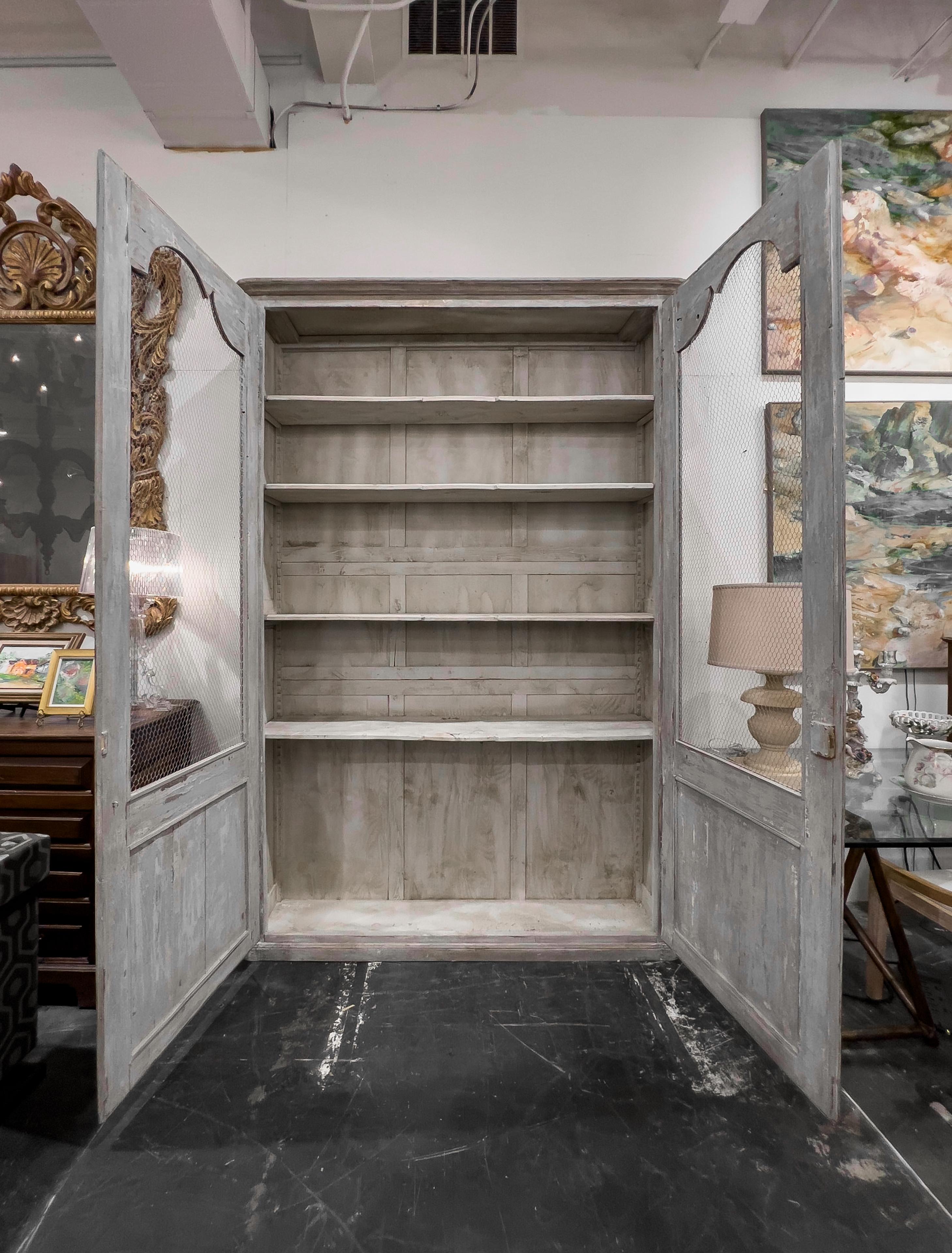 Directoire Style Painted French bookcase with Chicken wire doors. The exterior of the cabinet looks and has the construction of a 19th century piece yet the scalloped shelves and back panels look to be added at a later date. This piece is very