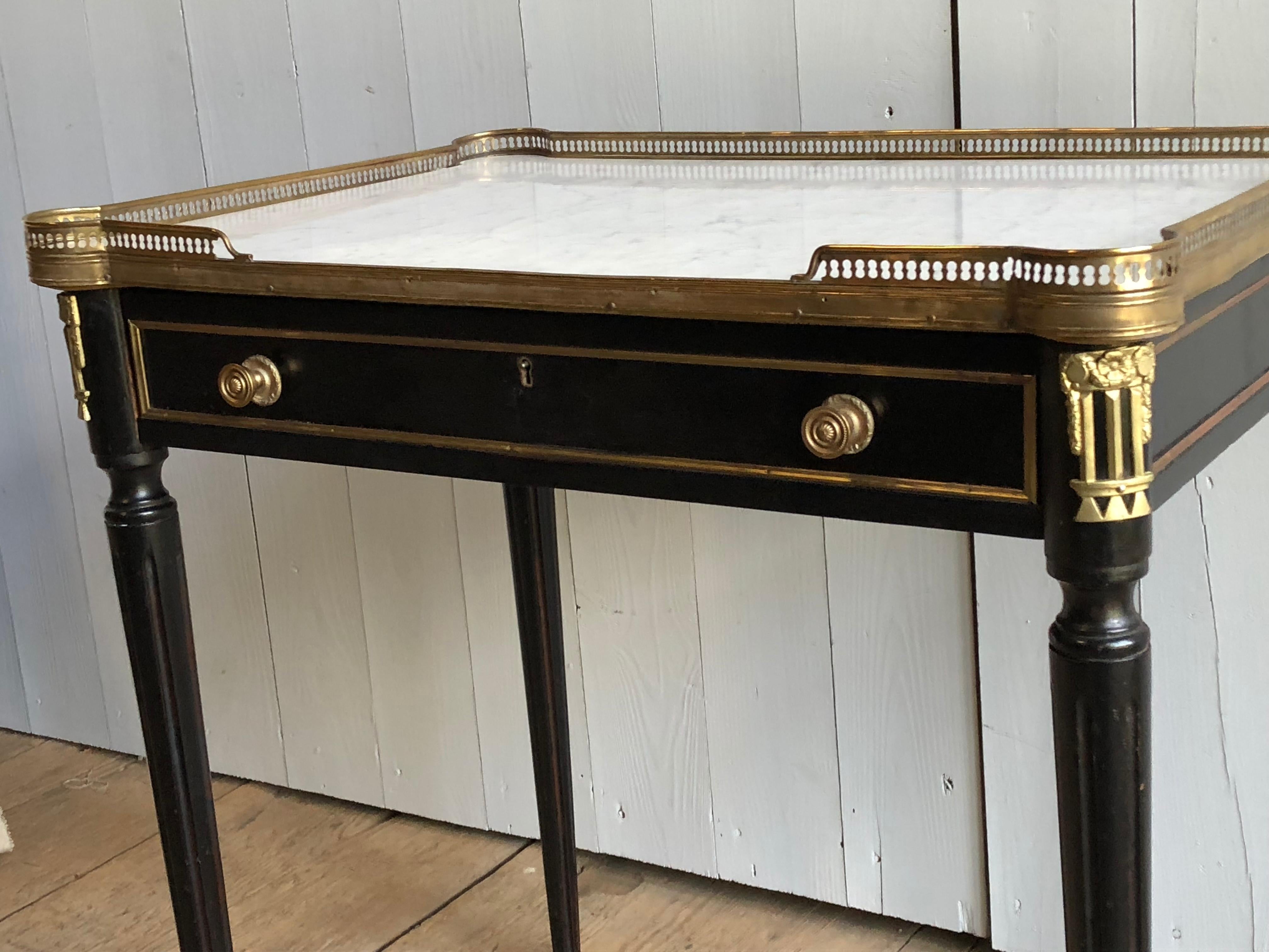 A French Directoire style writing or side table in black lacquer with a white marble top and brass mounts including a reticulated brass gallery, brass trim and foot caps, circa 1940. One long drawer with brass pulls over tapered and fluted legs.