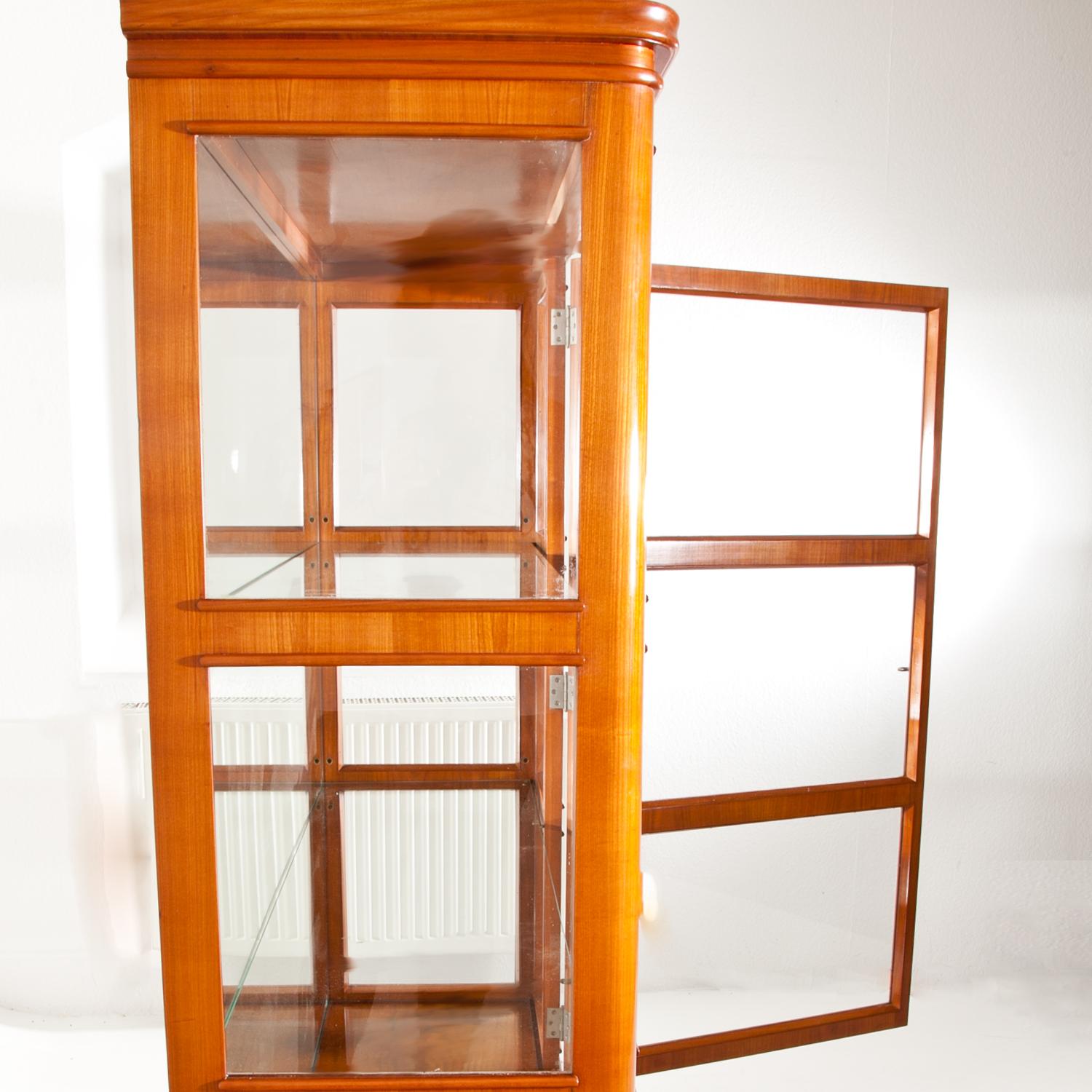 Large cherrywood display cabinet, standing on tall tapered feet. The display cabinet is glazed on three sides and the rear wall is mirrored. The door shows decorative inlays in the shape of banners.