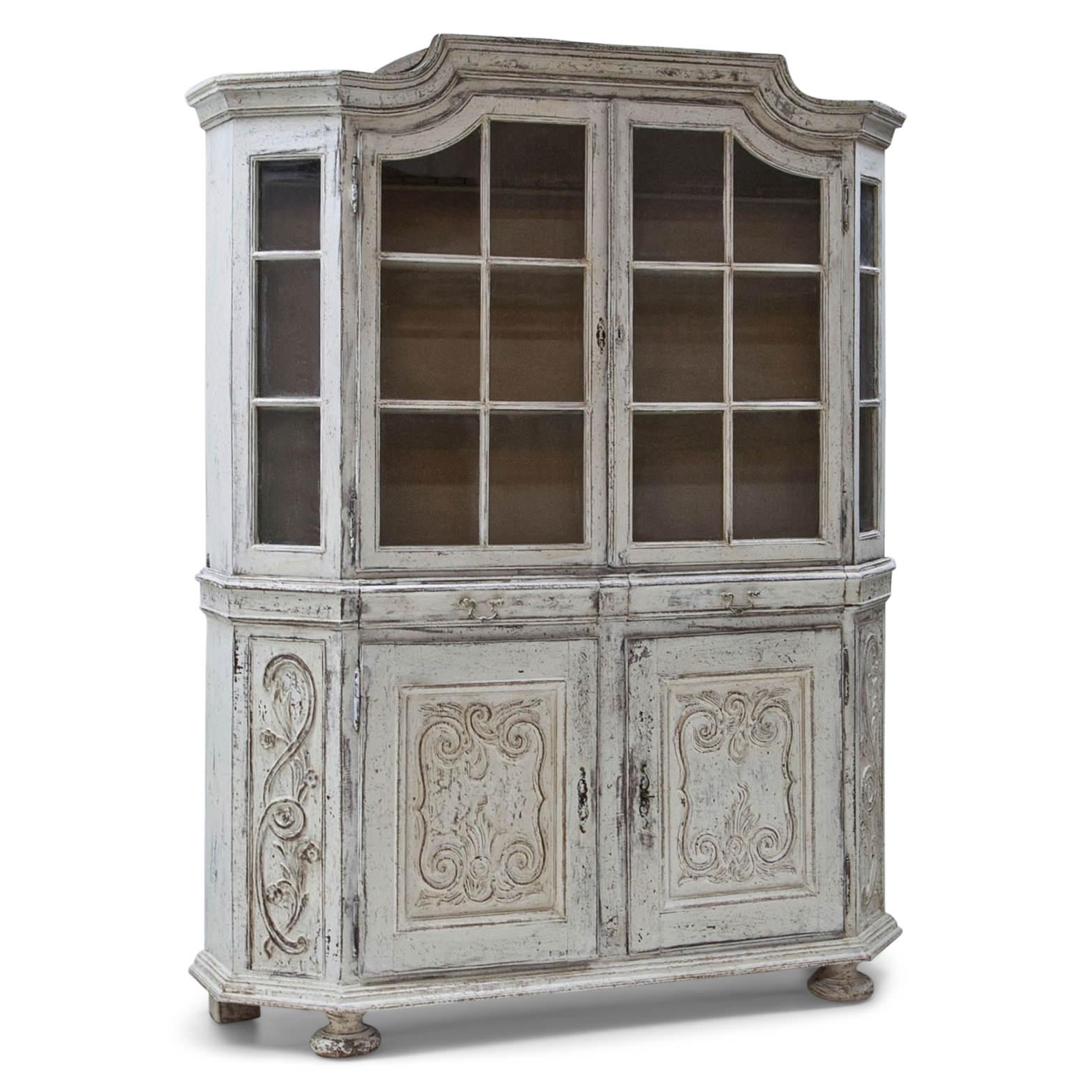 Large display cabinet with a two-door cabinet base, two drawers and a display case top part with two doors and strutted windows. The cornice slightly rises towards the middle and the corners of the cabinet are slanted. The white paintjob is new and