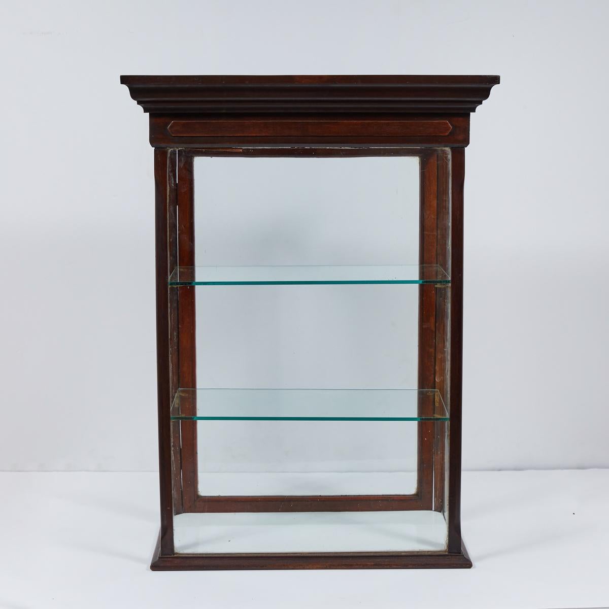 Tabletop display case or with glass walls and shelves and deep cherry-toned wooden siding from 1880s England. The beveled cornice gives the piece an elevated, architectural finish. Because there is glass on all sides, the piece can be displayed as a