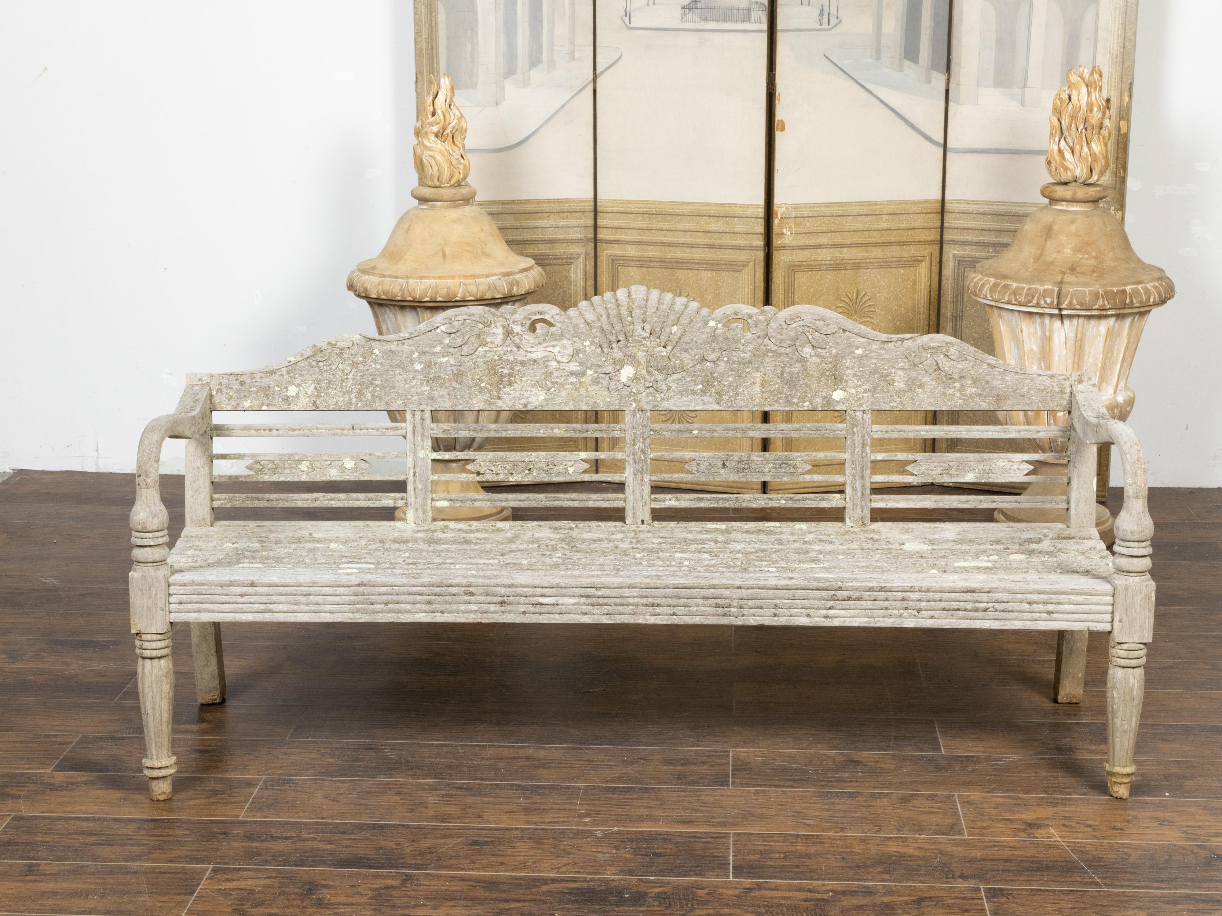A vintage English wooden garden bench from the mid 20th century, with light paint, carved foliage and fan motifs, distressed patina and scrolling arms. Created in England during the Midcentury period, this wooden bench features a slightly slanted