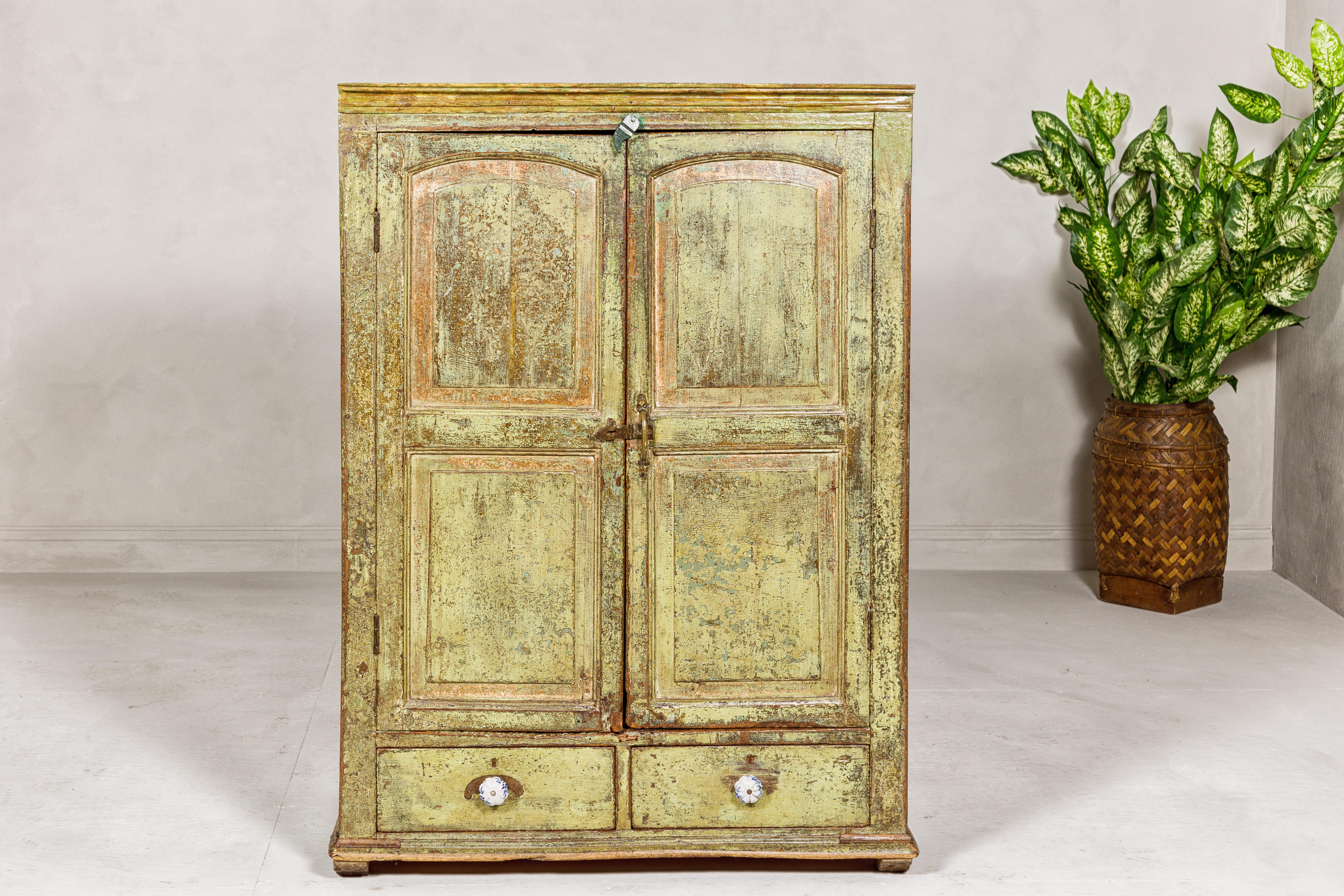 A small distressed green painted Indian cabinet from the 19th century with paneled doors and two lower drawers. This 19th-century Indian cabinet exudes rustic elegance with its distressed green paint finish, lending an air of historic charm to any