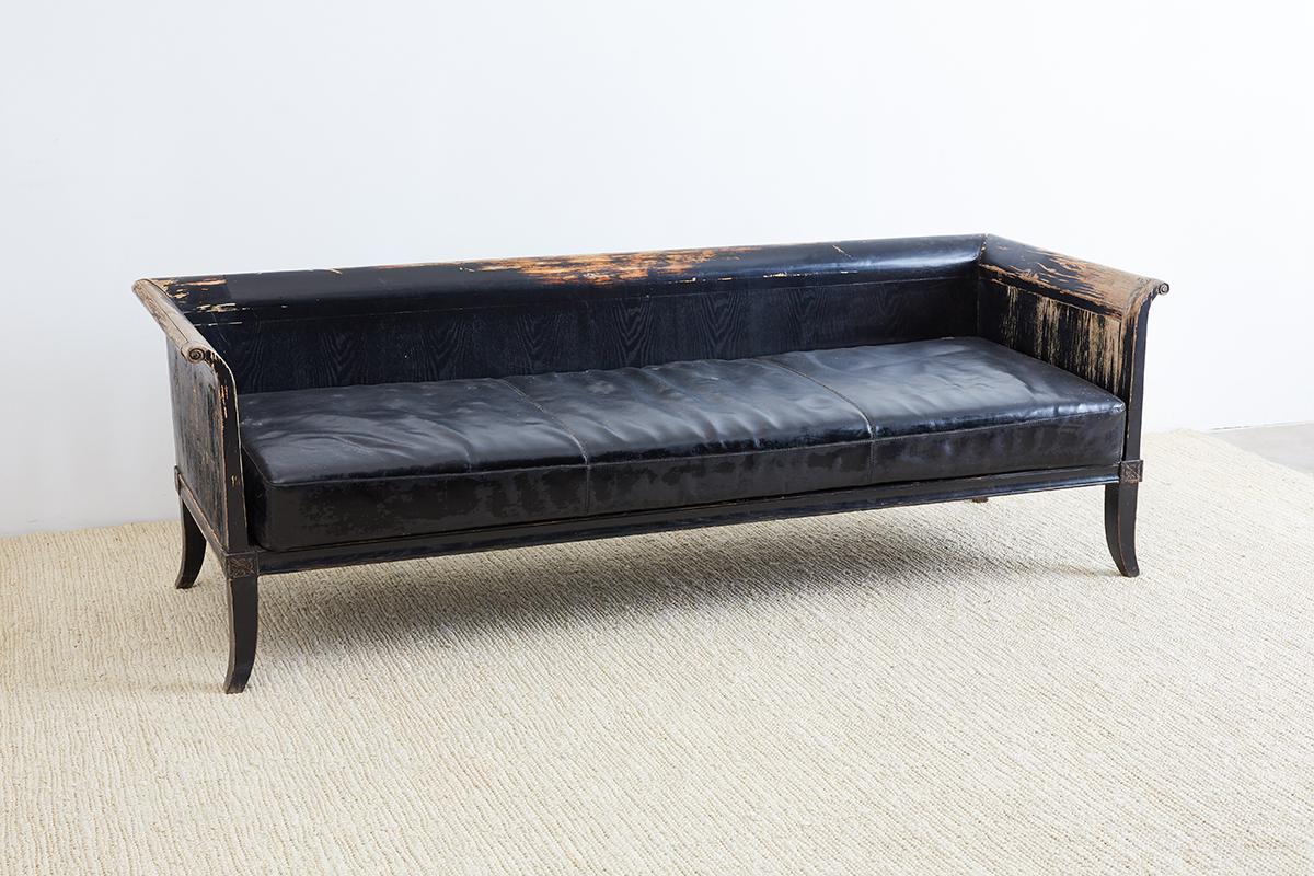 Dramatic neoclassical Gustavian style sofa or wooden bench featuring a worn, weathered, and chipped black lacquer wood finish. Scrolled arms and back with a sleek low profile. Faux leather naugahyde loose cushion with vintage worn leather patina
