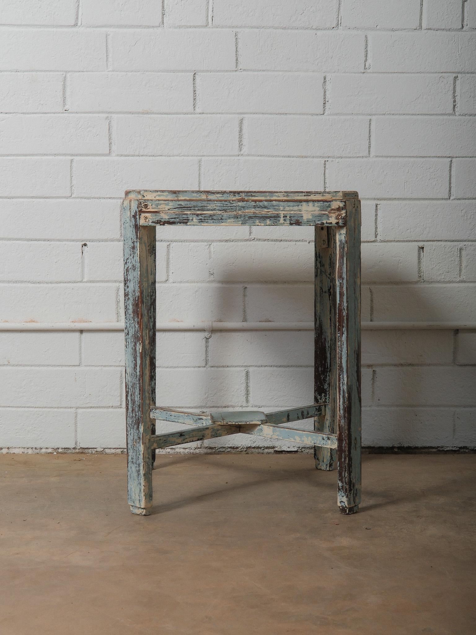 This distressed painted wooden accent table features a beautifully weathered blue finish that highlights years of natural patina and character. Its compact square form, subtly rounded edges, and crossed stretcher base give it both charm and