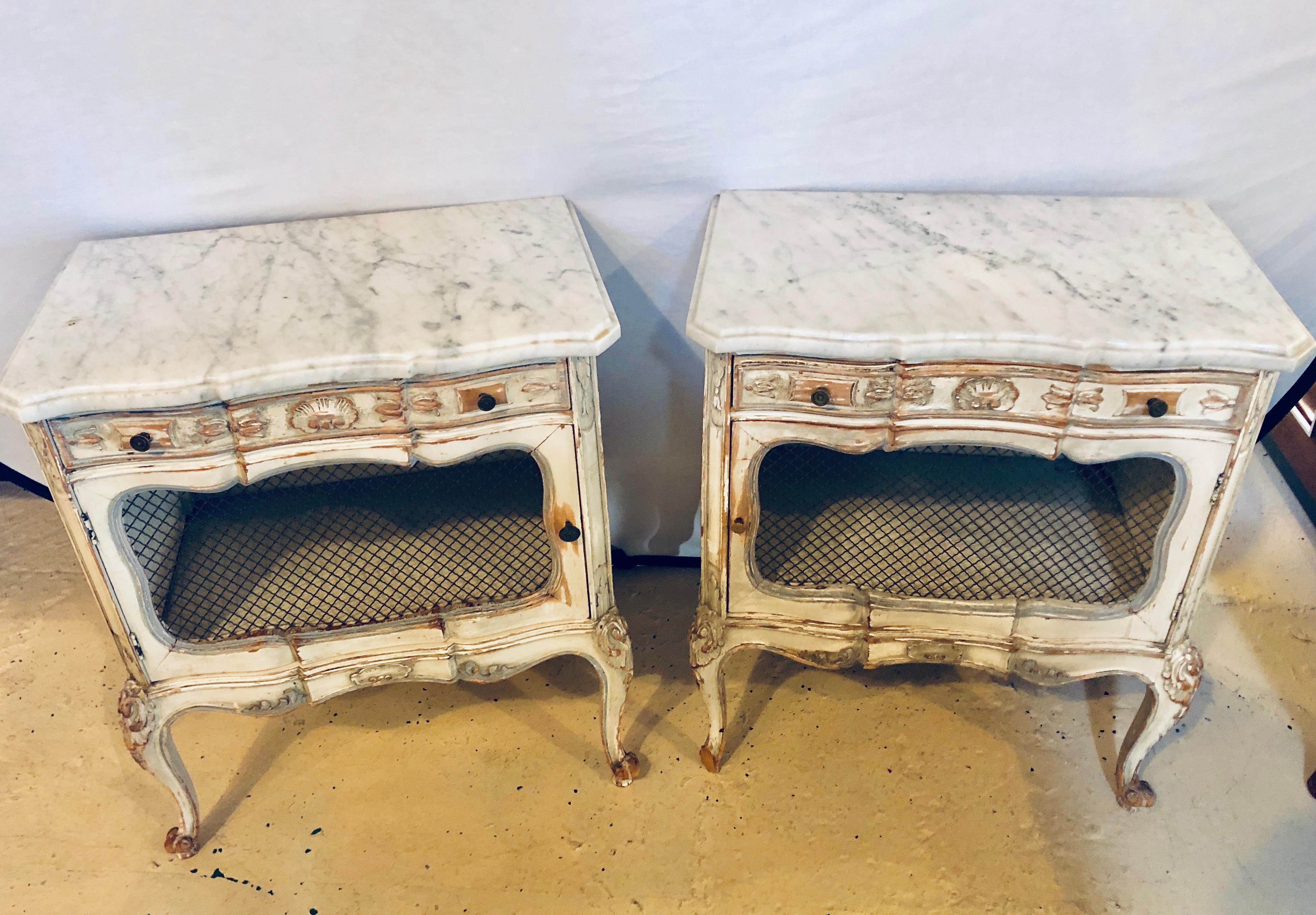 A distressed pair of marble top end tables or bed/night side stand. These wonderfully decorative parcel paint and gilt designed end tables each have a marble top over a case with one single drawer and a pair of lower grilled doors leading into a