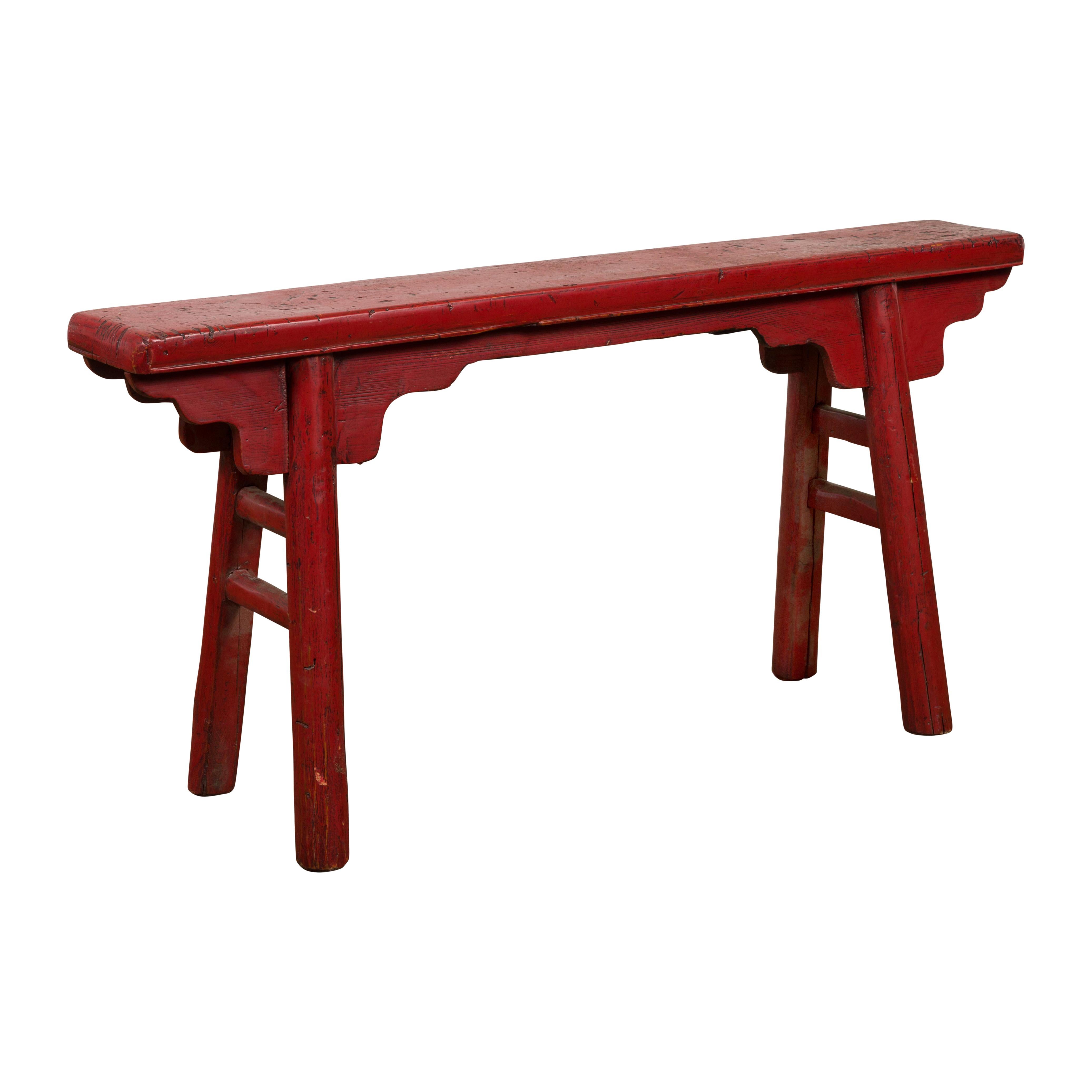 A Chinese vintage Ming Dynasty style red lacquer wooden bench from the mid 20th century with A-form base, carved spandrels and distressed finish. Embrace the timeless allure of this Chinese vintage Ming Dynasty-style red lacquer wooden bench from