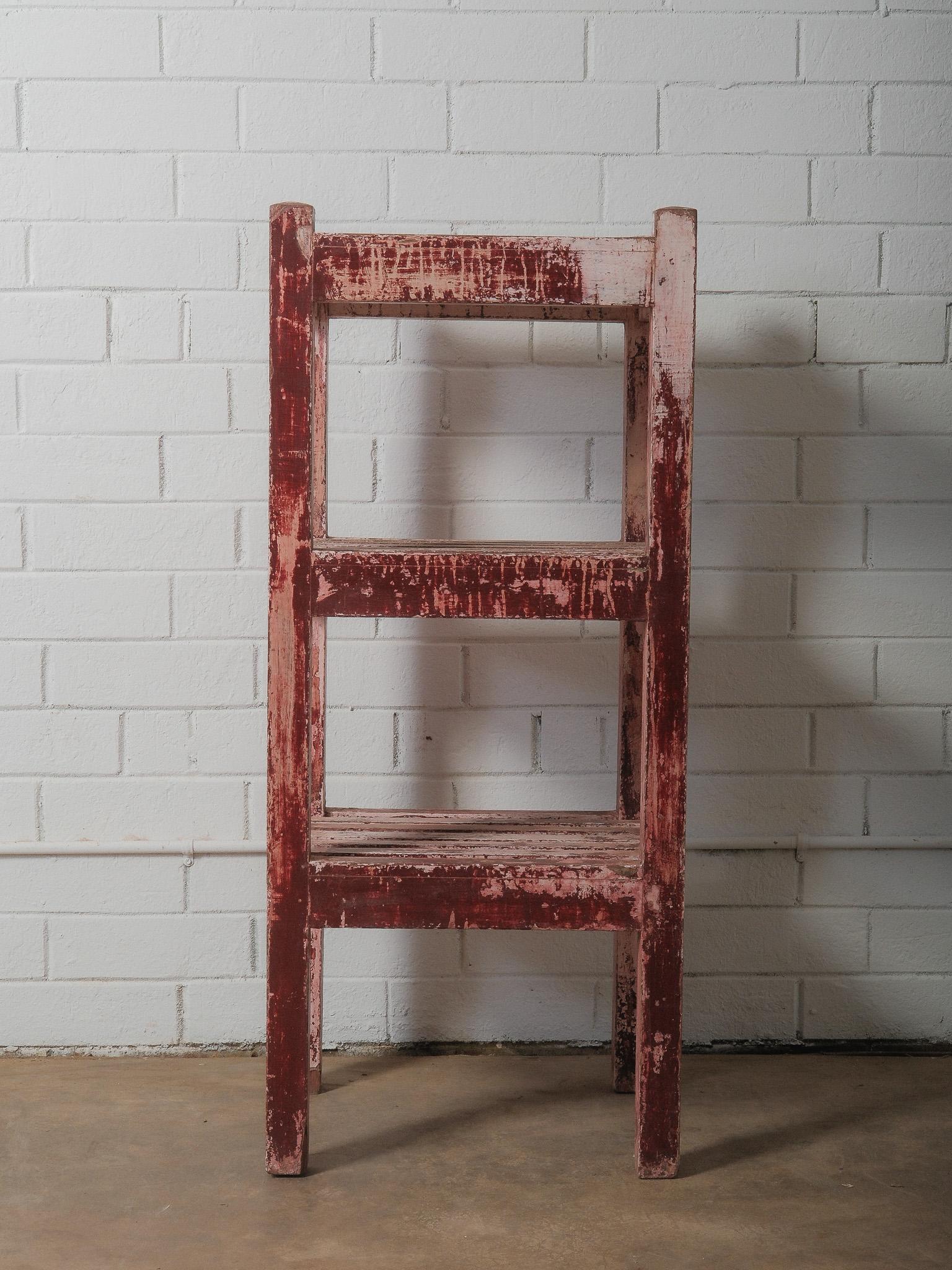 A sculptural wooden shelf whose presence is defined by an extraordinary, time-worn surface of deep red and chalky pink paint. The finish has softened through decades of use, creating an organic pattern of abrasion, exposed grain, and mineral-like