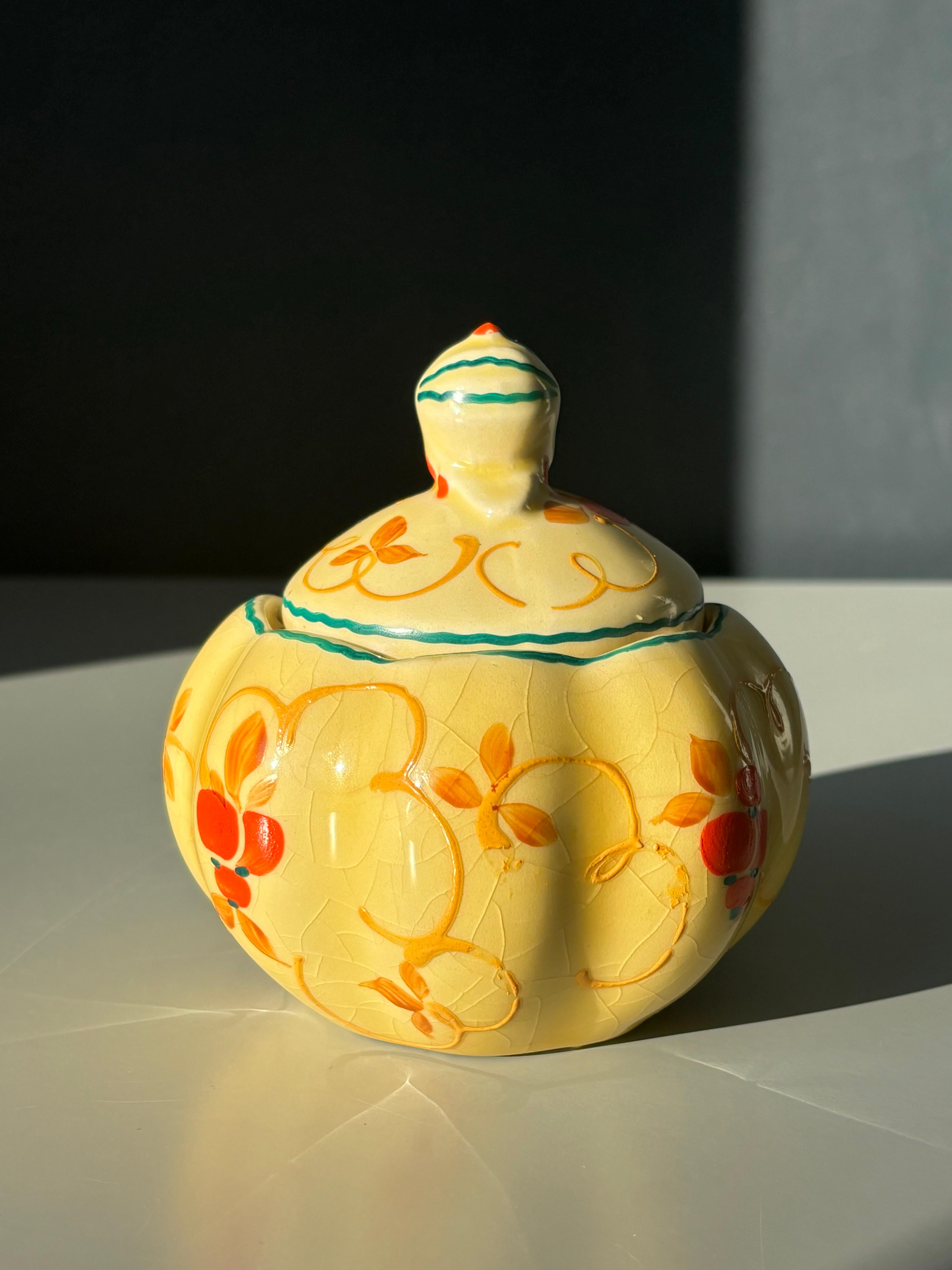 Colorido cuenco bohemio art nouveau con tapa y pequeña hendidura para una cuchara. Decoraciones de bayas pintadas a mano en naranja brillante sobre un cálido esmalte amarillo y líneas florales orgánicas en verde y caramelo dorado.
Diseñado y