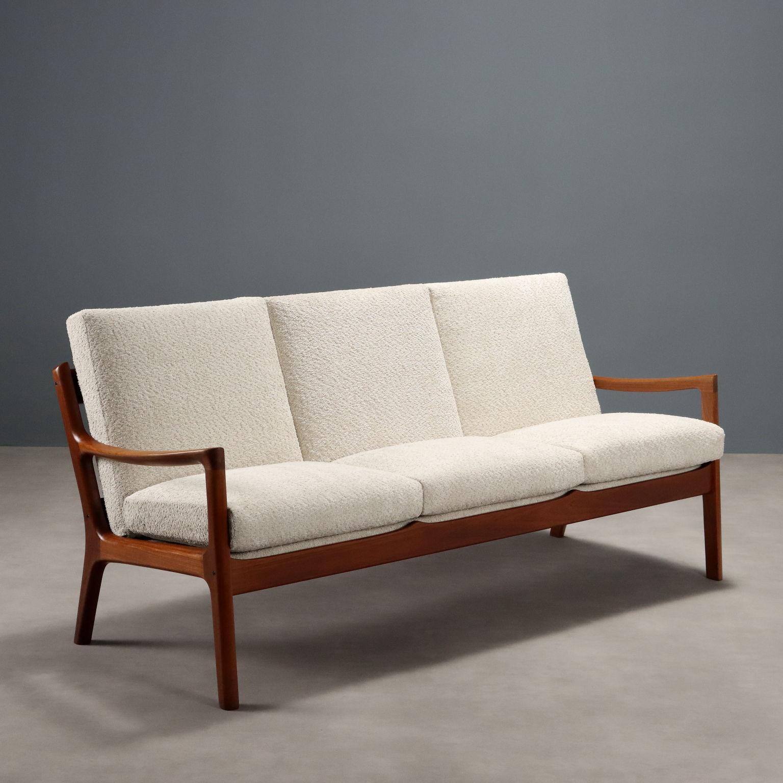 Divano vintage a tre posti con struttura in legno di teak a vista, seduta e schienale con imbottitura in espanso e molle, tappezzeria nuova in tessuto bianco. Presente il marchio della manifattura.