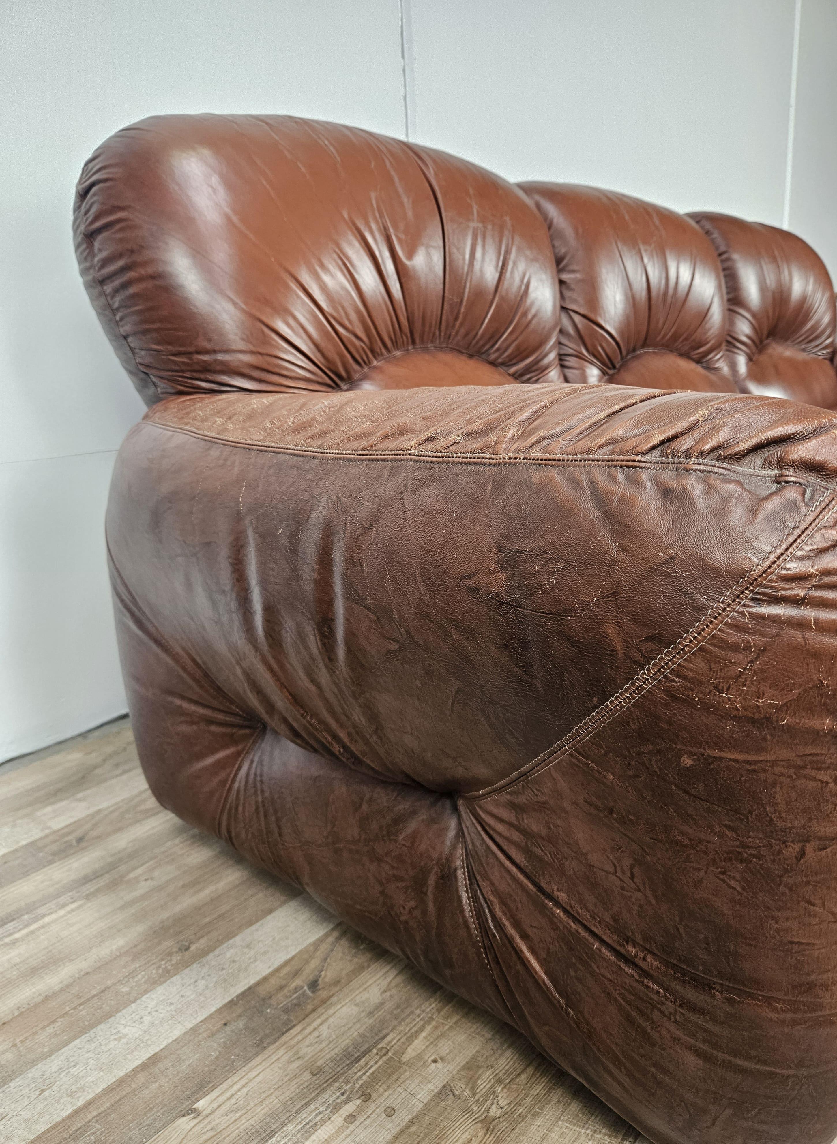 Bequemes 1970er-Jahre-Sofa aus braunem Leder mit Polstern und schwarzen Kunststofffüßen.

Sitzhöhe ca. 46 cm.

Es zeigt Alters- und Gebrauchsspuren wie auf dem Foto zu sehen, das Leder ist stellenweise abgenutzt.
Soweit möglich, wurde es mit einer