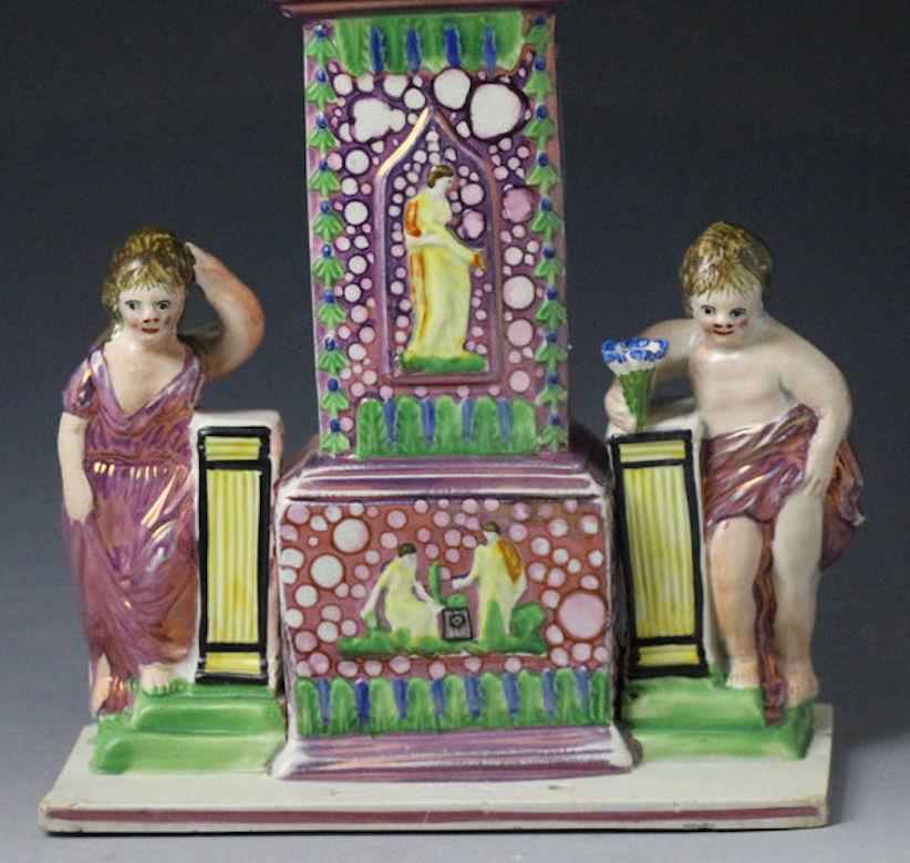 A large and rare pottery version of a Sunderland Pottery watch stand in the form of a long case clock flanked by cherubs who are modeled standing by plinths. The piece decorated with excellent quality pink splash luster and enamels. The base of the