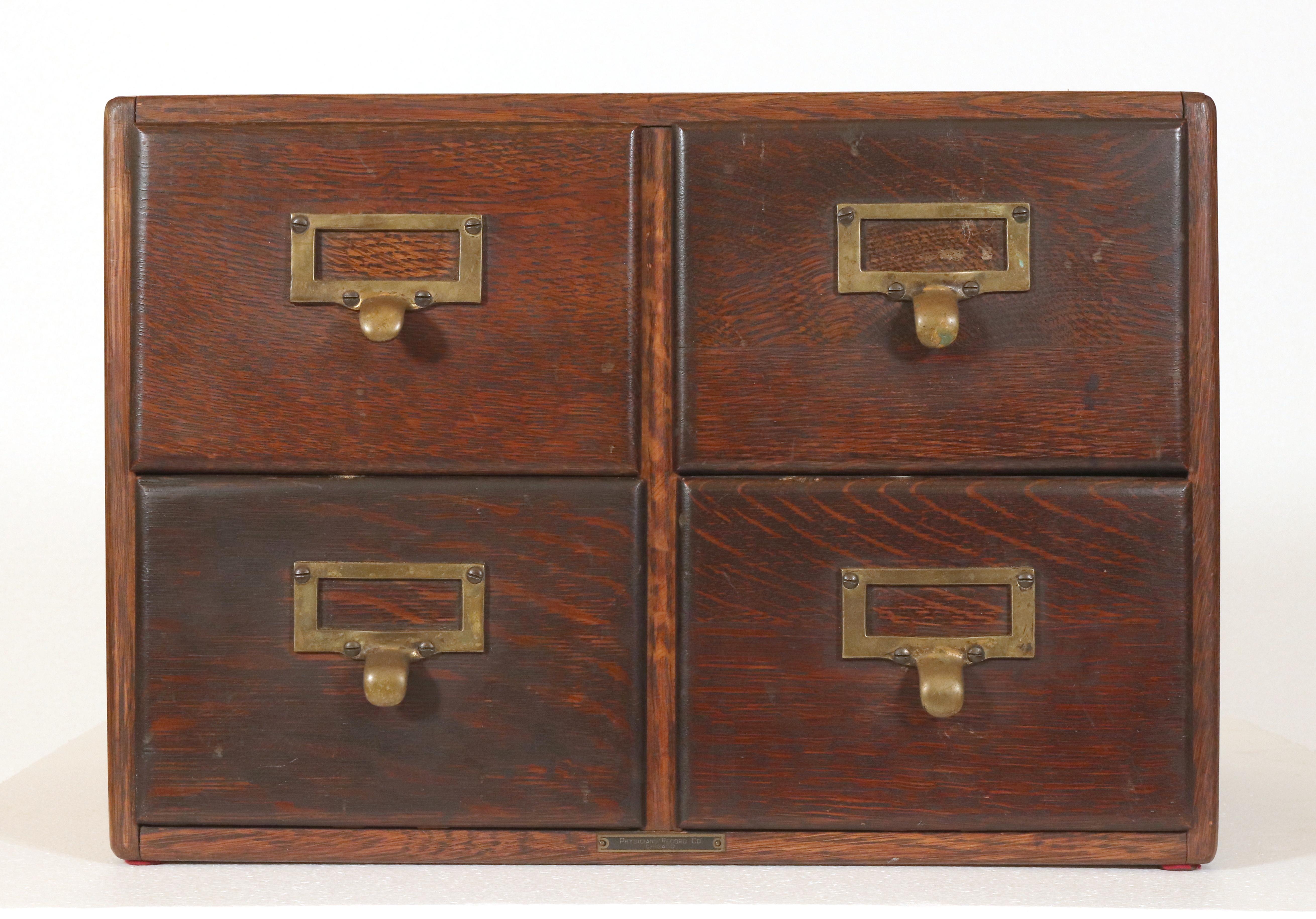 Antique tabletop doctor's card catalog made of dark stained quarter sawn oak. It features four dovetailed drawers with brass label slot pulls. On the center bottom, there is a plaque inscribed 
