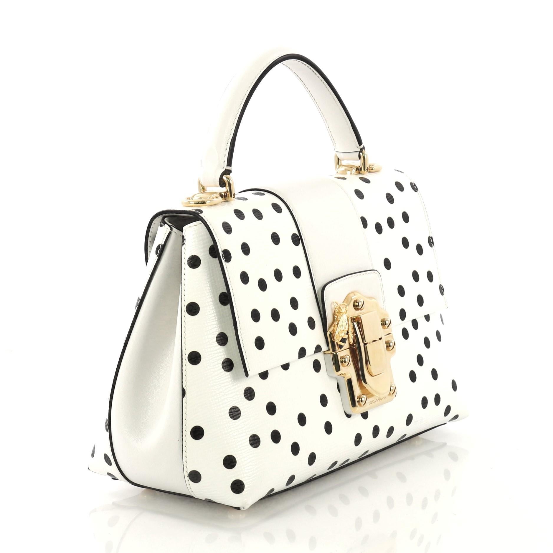 This Dolce & Gabbana Lucia Top Handle Bag Printed Leather Medium, crafted from black and white polka dot printed leather, features a leather top handle, adjustable shoulder strap, push-lock fastening with bee detailing, protective base studs, and