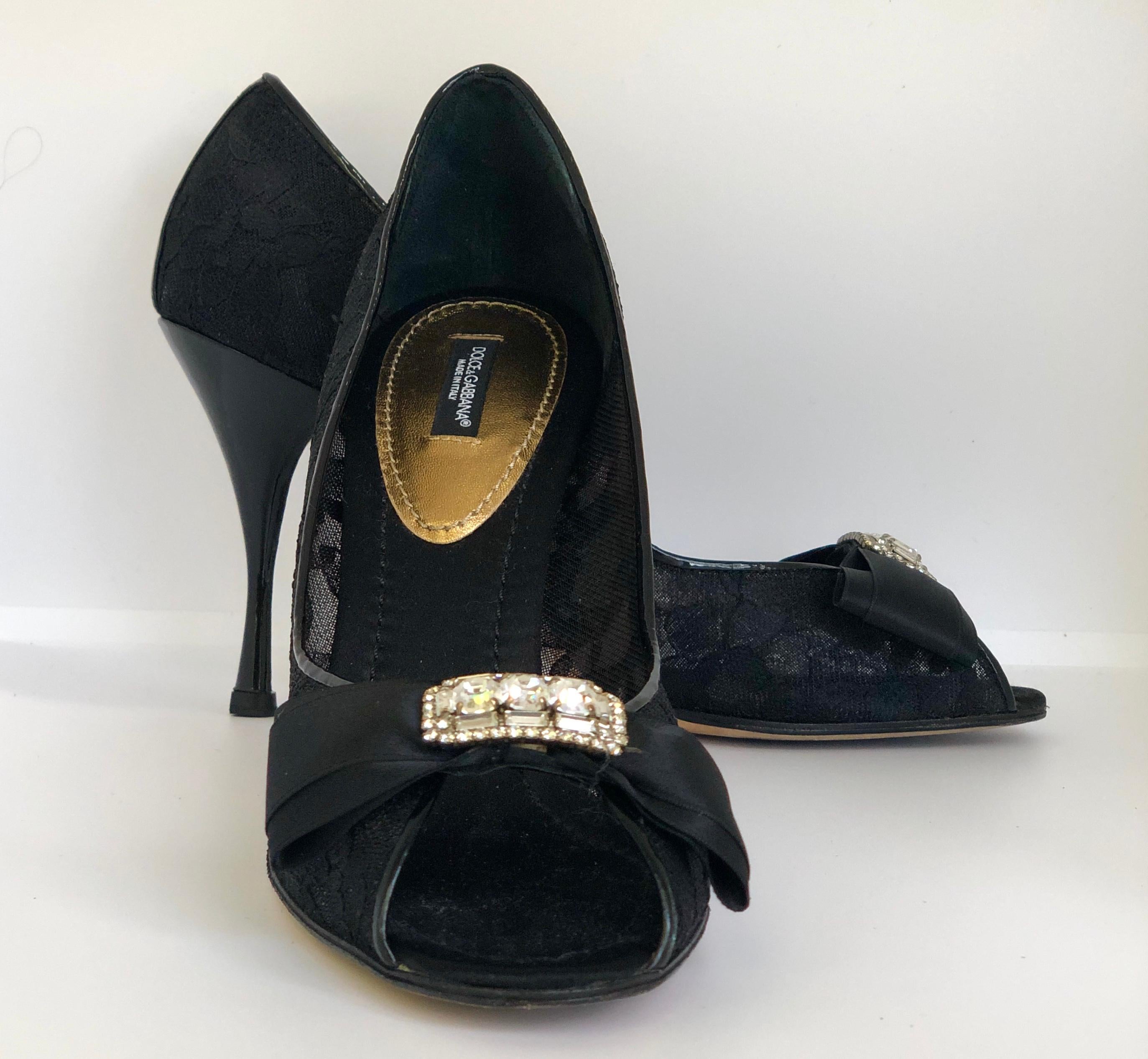 Offered is a pair of Dolce & Gabbana jewel embellished satin bow with black lace and patent leather peep toe high heel pumps / shoes

Make:  Dolce & Gabbana
Place of Manufacture:  Italy
Color:  Black
Materials:  Patent leather, satin silk, lace and
