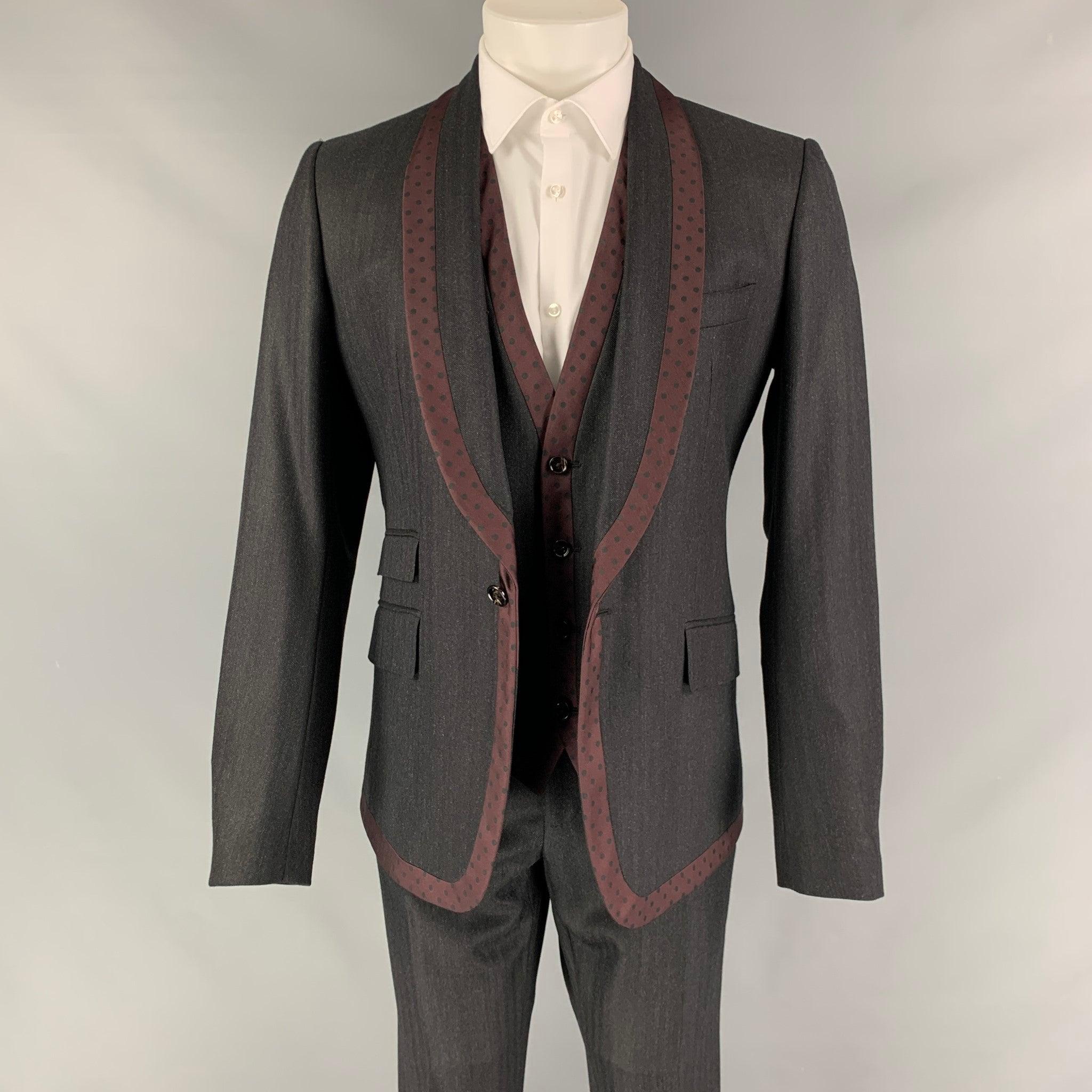 Le costume 3 pièces DOLCE & GABBANA est en laine vierge à pois gris et bordeaux, avec doublure intégrale. Il comprend un manteau sport à un seul boutonnage et col châle, un gilet assorti et un pantalon à devant plat. La taille et la longueur des
