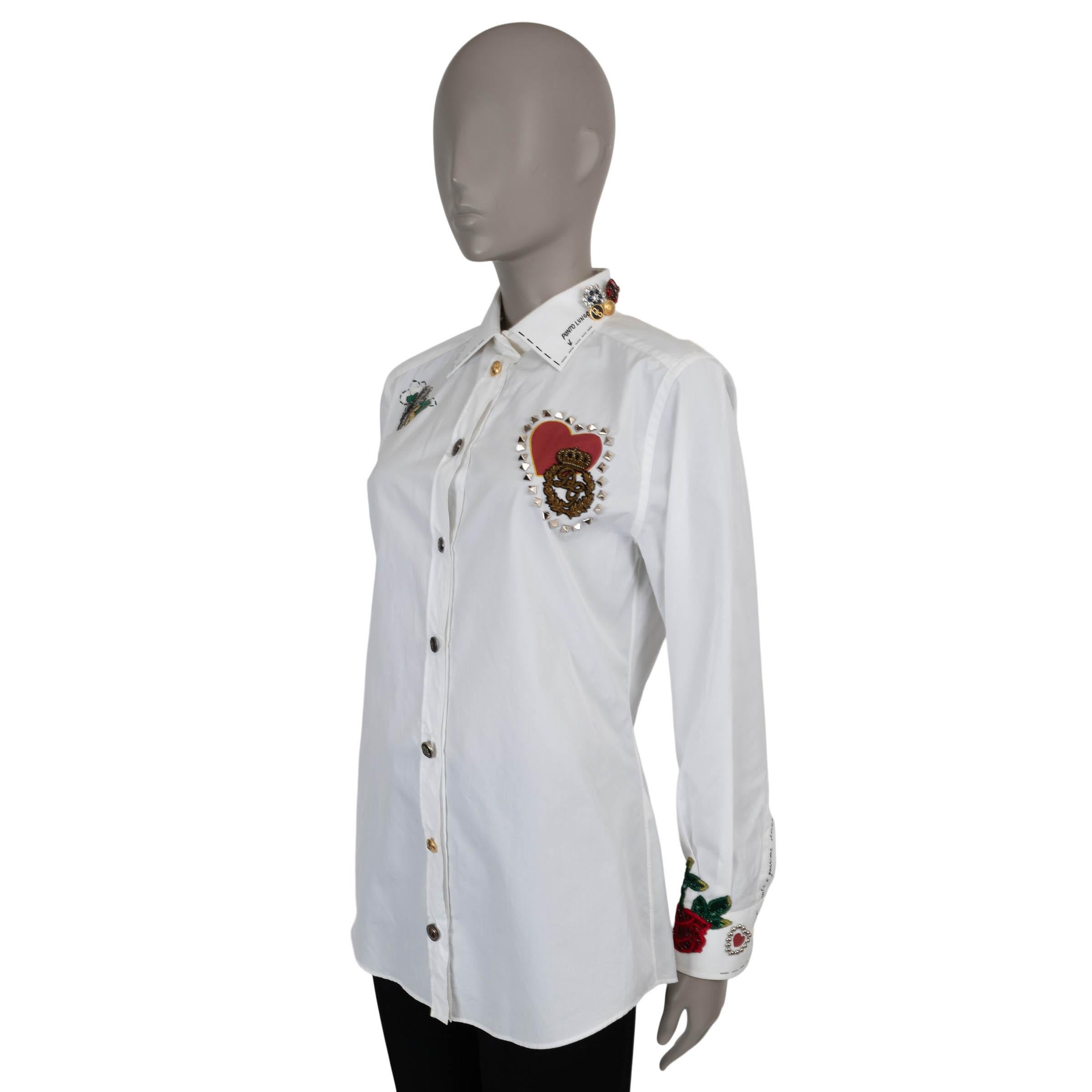 This 100% authentic Dolce & Gabbana button-up shirt in white poplin cotton (100%) is a statement piece adorned with intricate embellishments. Featuring a red printed heart and an embroidered DG crown outlined in silver metal pyramid studs on the