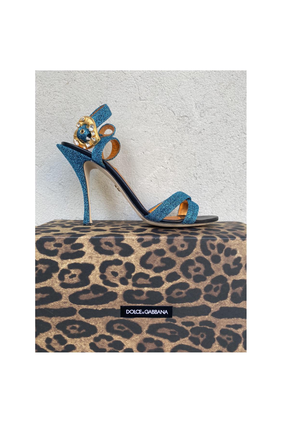 Sandali Dolce & Gabbana in lurex azzurro con cinturino alla caviglia e fibbia a forma di cuore. Numero 38. Ottime condizioni, solo una tacchetta, come si può vedere dalle foto, è rotta e deve essere sostituita. Con scatola originale.