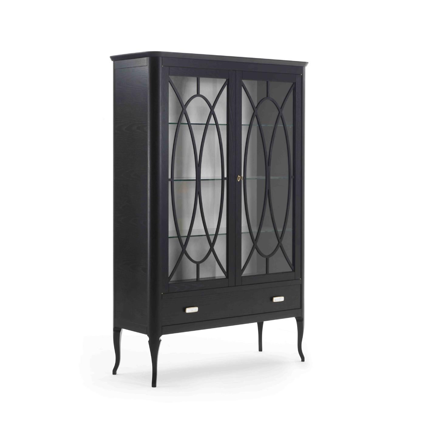 Fashioned of wood with ash veneer and ebonized finish, this refined display cabinet features a rectangular frame with cabriole legs and two glass doors accented with an open design of intersecting ovals and a single oval brass knob. The interior has